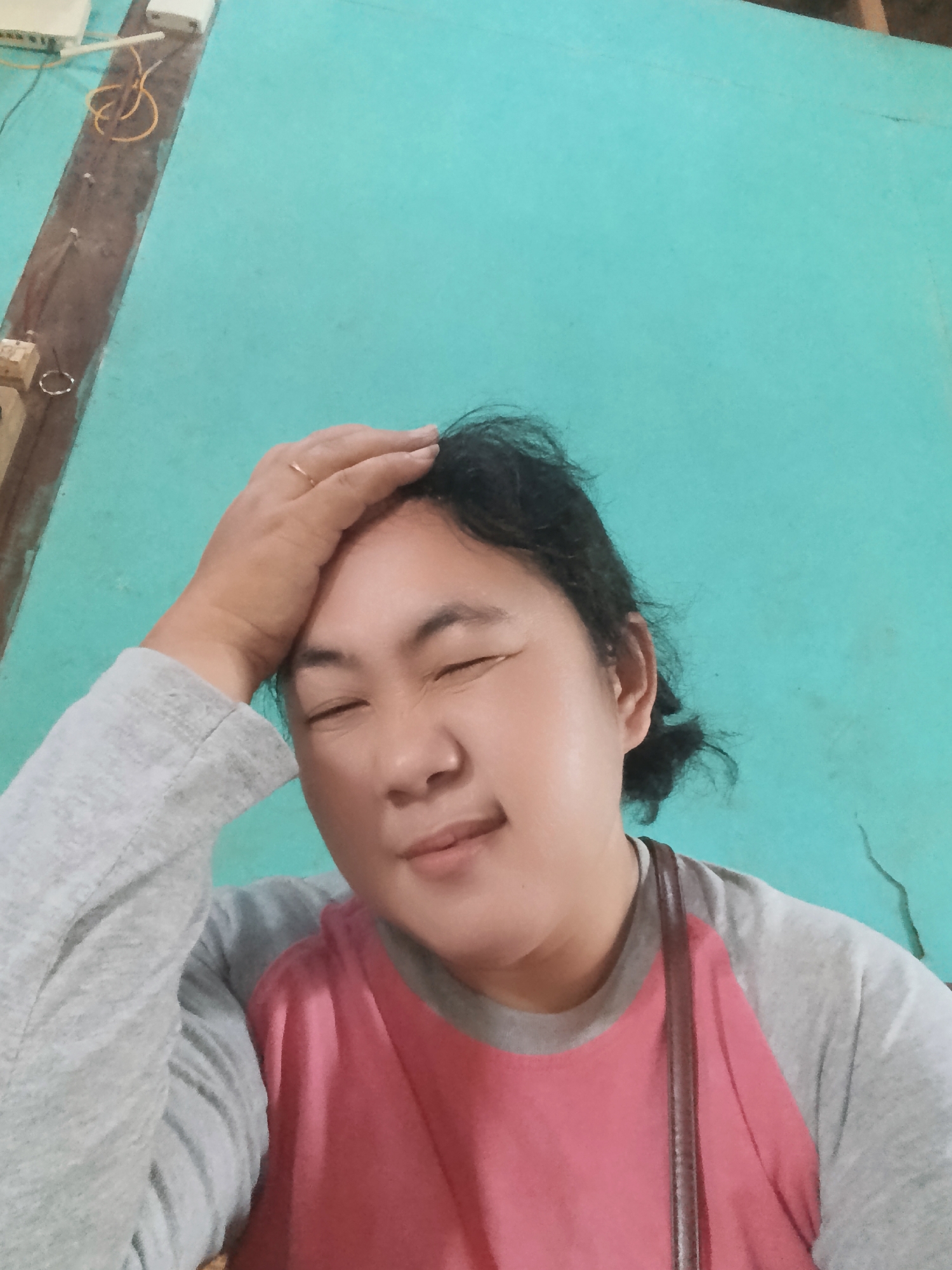 A medium shot captures a woman with her eyes closed and her right hand resting on her forehead, suggesting discomfort or contemplation. She has dark hair styled in a bun and is wearing a grey long-sleeved shirt with a pink yoke. The background is a solid, turquoise-colored wall. The overall image appears to be a selfie.