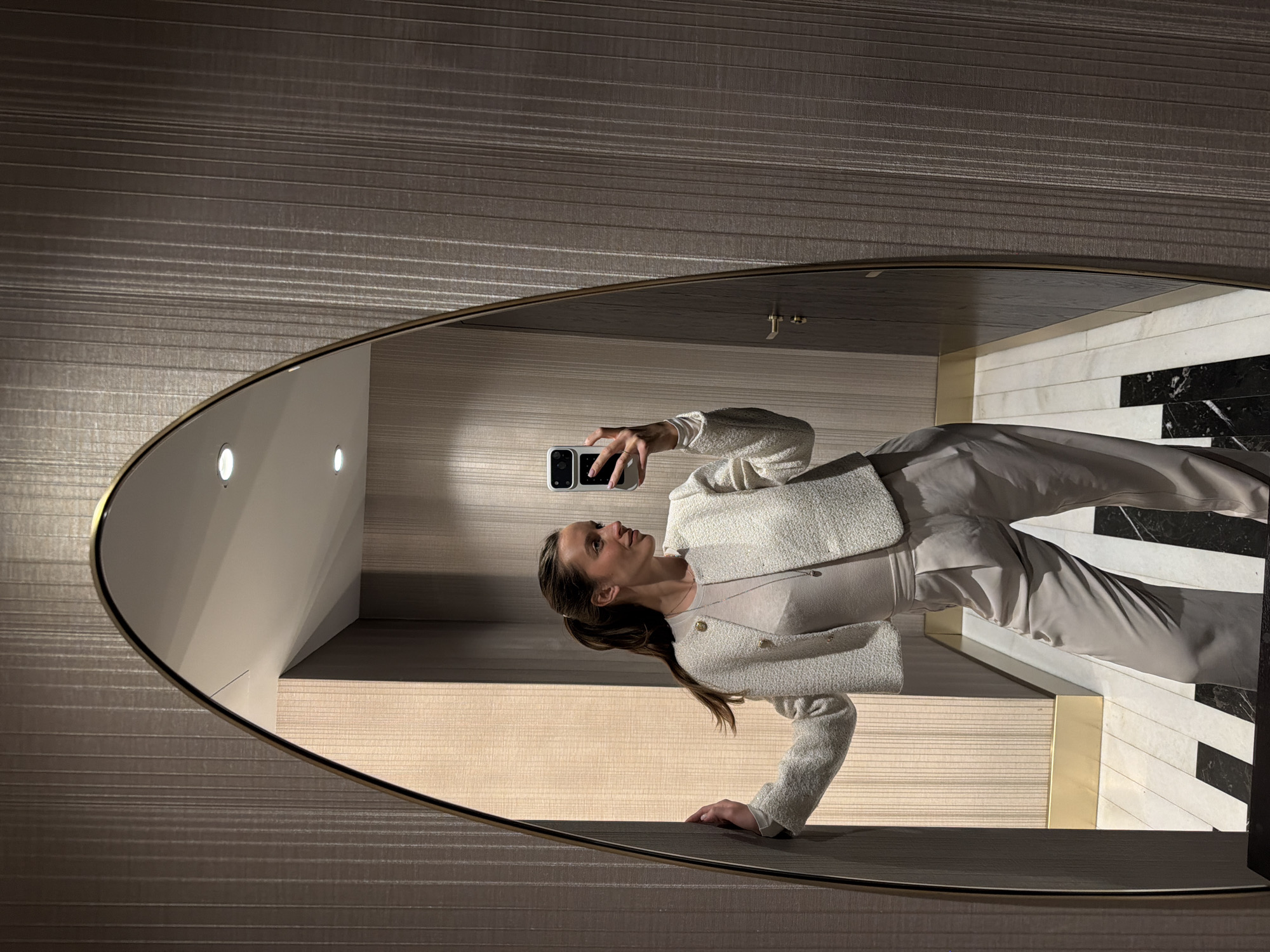 An upright, full-length mirror selfie captures a woman in an elegant indoor setting, likely in Budva, Montenegro. The woman, with long brown hair tied back, is seen from the chest up, looking slightly upwards and to her left, not directly at the smartphone in her right hand. She is dressed in a sophisticated ensemble featuring a light-colored, possibly off-white or cream, textured blazer with gold buttons, a matching top, and light-toned trousers. A delicate necklace with a small pendant is visible around her neck.
She holds a white smartphone, aimed to capture her reflection in the large, oval-shaped mirror, which has a thin, dark frame. The wall behind the mirror is covered in a dark grey-brown wallpaper with subtle horizontal stripes. The mirror's reflection reveals more of the interior: a distinct floor pattern composed of alternating stripes of light (possibly white or light beige) and dark (black marble-like) tiles, interspersed with horizontal gold-toned accents. Recessed lighting fixtures are visible on the ceiling within the reflection, alongside light-colored walls. A brass-colored tap is also faintly reflected, suggesting the setting might be a luxurious restroom or vanity area. The lighting is artificial and bright, emphasizing the clean lines and upscale materials of the environment. No text is visible in the image.