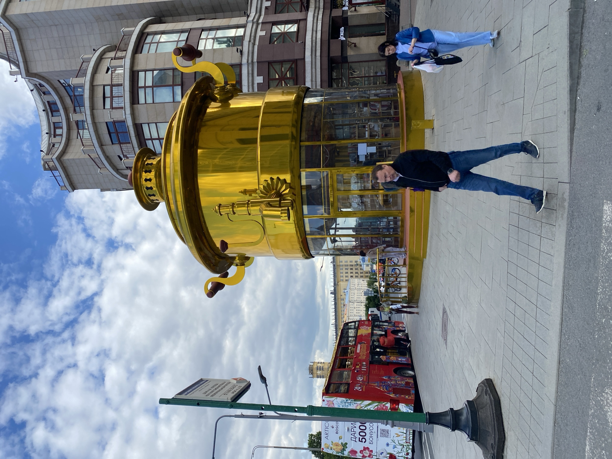 The image captures a street scene in Moscow, Russia, featuring a large, golden, samovar-shaped building. A man in a black jacket and jeans stands near the structure. Another woman in a light blue suit is seen walking near the man. A red double-decker tour bus is positioned behind the samovar. Street signage is partially visible in the foreground. The architectural backdrop includes buildings with light facades and dark-framed windows. The sky overhead is partly cloudy.