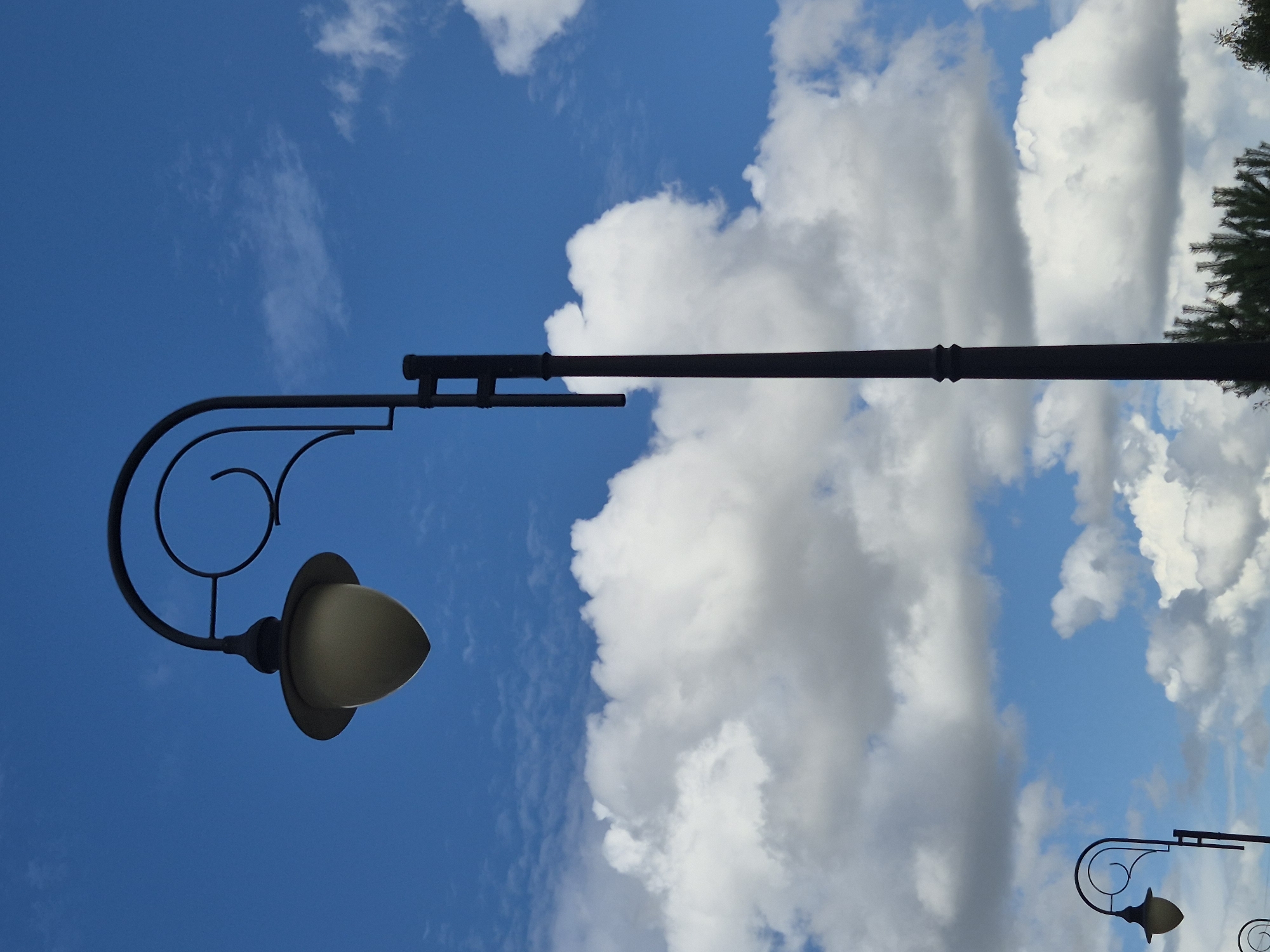 The image displays a tall, dark lamppost against a bright blue sky with scattered white clouds. The lamppost has an ornate curved arm supporting a cream-colored lamp. In the background, the tops of evergreen trees are visible. The image appears to be taken from a low angle, looking up at the lamppost and the sky. There are no people or animals visible in the frame. The setting is outdoors, possibly in a park or along a street. The image does not provide any specific details that would indicate its location in the city of Kokshetau, Kazakhstan.