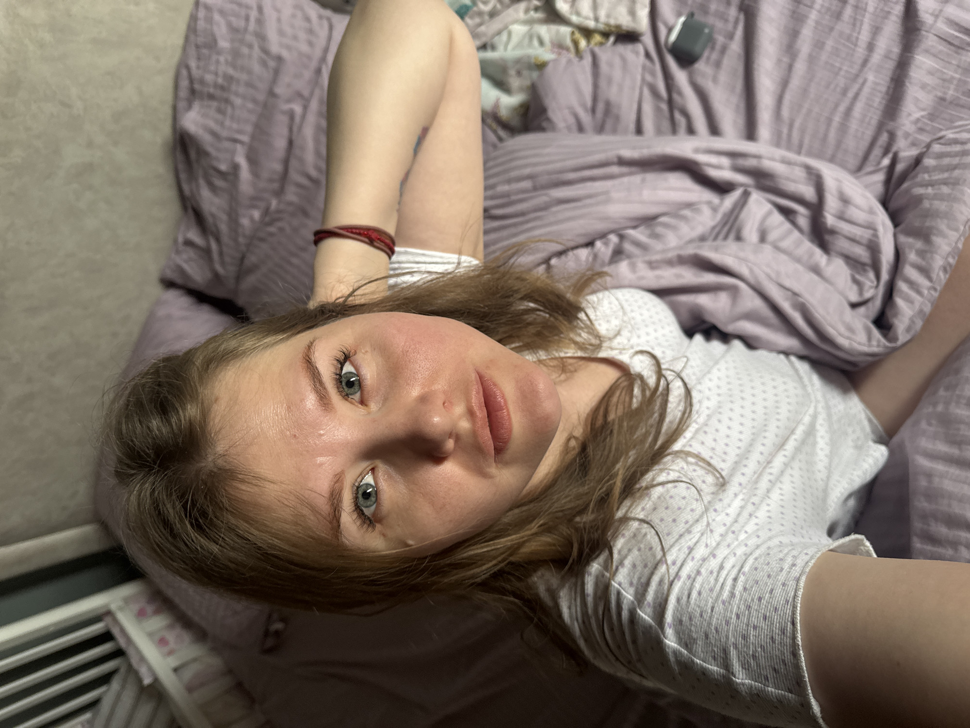 A woman is lying on a bed with lavender-colored bedding. She is wearing a white, patterned top and has a red bracelet on her left wrist. Her light brown hair is disheveled. She has fair skin, light blue eyes and a slightly flushed face. A white radiator is visible in the lower left corner of the frame. A small, dark object, possibly a case for earbuds, is on the bed near her head. The scene appears to be indoors. The image lacks location-specific features that could indicate Gornyi (Dvurechenskoe S/P), Russia.