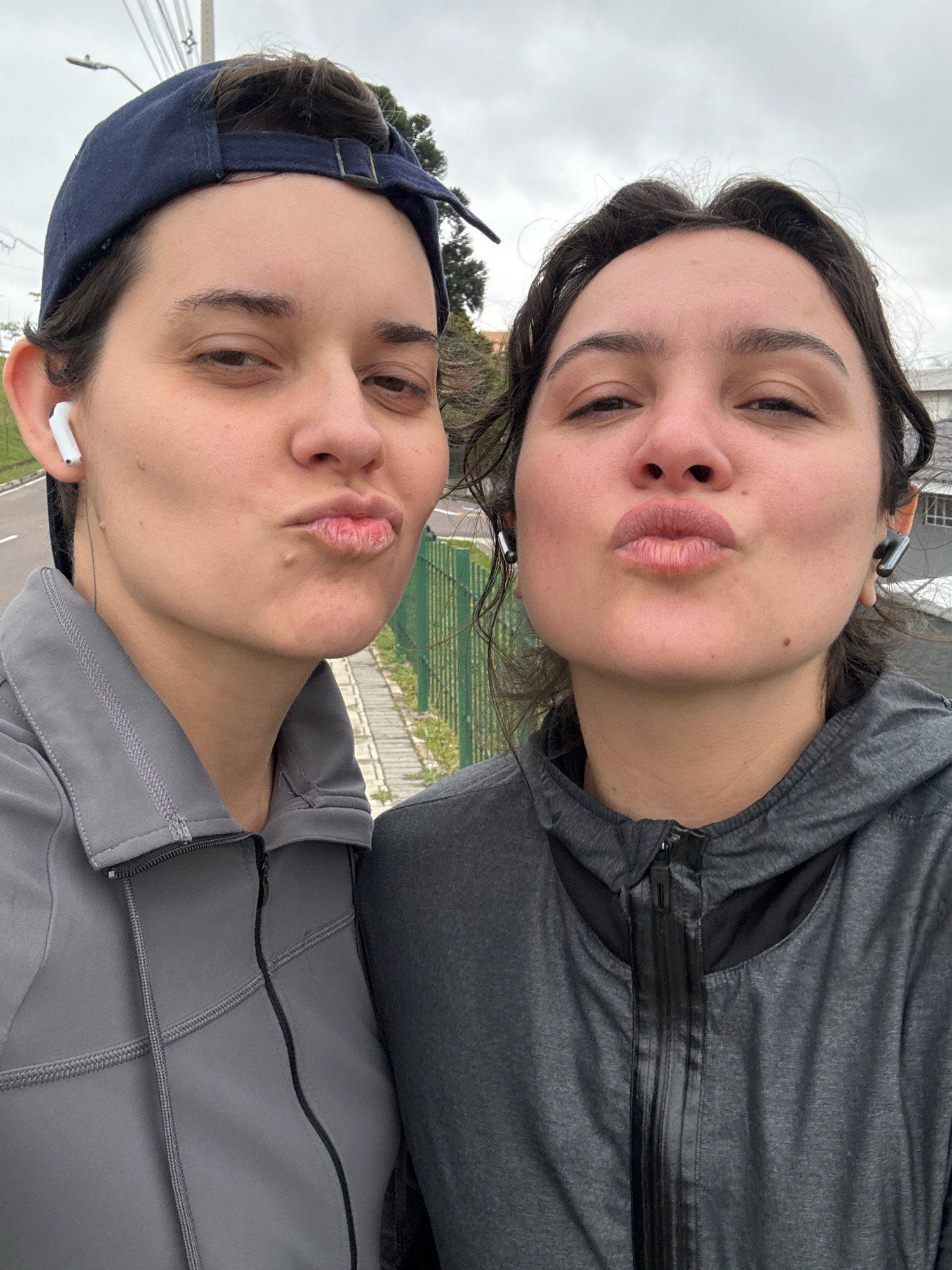 Two individuals, both with pursed lips as if to give a kiss, are taking a selfie outdoors. The person on the left wears a navy blue baseball cap and a grey zip-up jacket with visible stitching and a black cord. They have white earbuds in their left ear. The person on the right wears a dark grey hooded jacket with a prominent black zipper. Both individuals appear to be close together, suggesting they are friends or a couple. The background shows a paved path, a green metal fence, and trees, indicating an outdoor recreational area, possibly a park or a running track within the city of Curitiba, Brazil, given the context. The sky is overcast.
