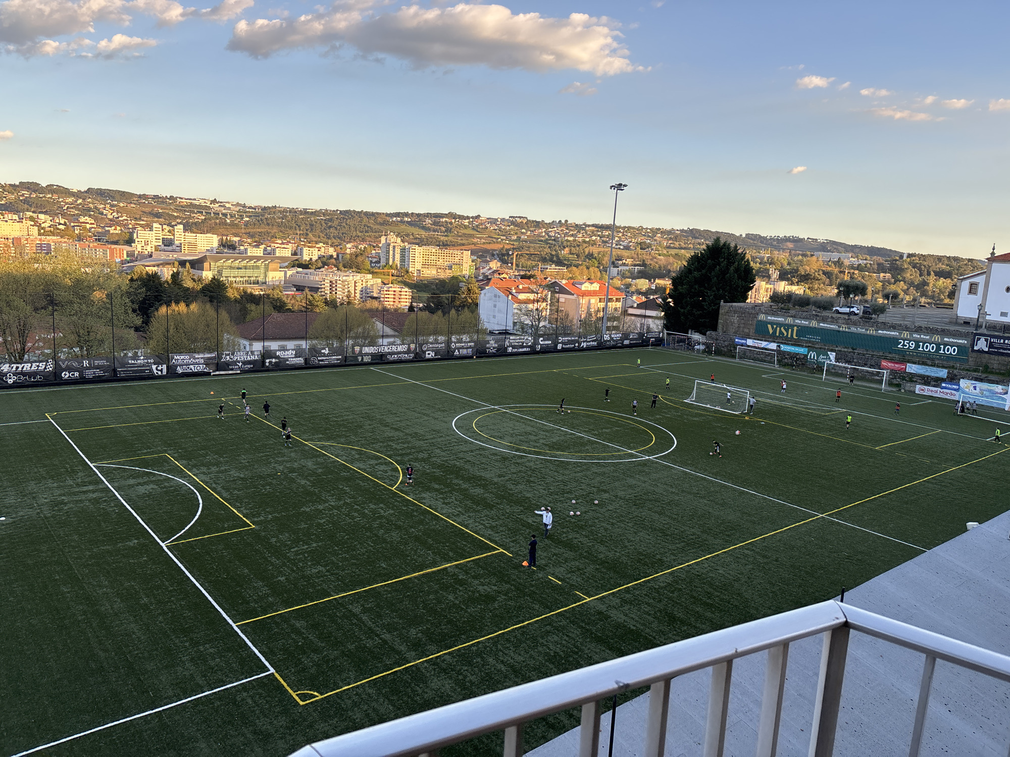 The image depicts a soccer field located in Vila Real, Portugal. The field is a synthetic turf pitch with white lines marking the boundaries and central circle, and yellow lines marking penalty areas and corners. Several people are visible on the field, engaged in what appears to be a practice session. Some individuals are actively running and kicking soccer balls, while others, including a coach, are standing still, presumably providing instruction. The field is surrounded by a fence adorned with commercial advertisements. Beyond the fence is a cityscape of Vila Real, featuring buildings of varying sizes and architectural styles. The sky is partly cloudy, indicating daytime, and the overall light suggests it might be late afternoon. A small portion of a balcony railing is visible in the foreground.