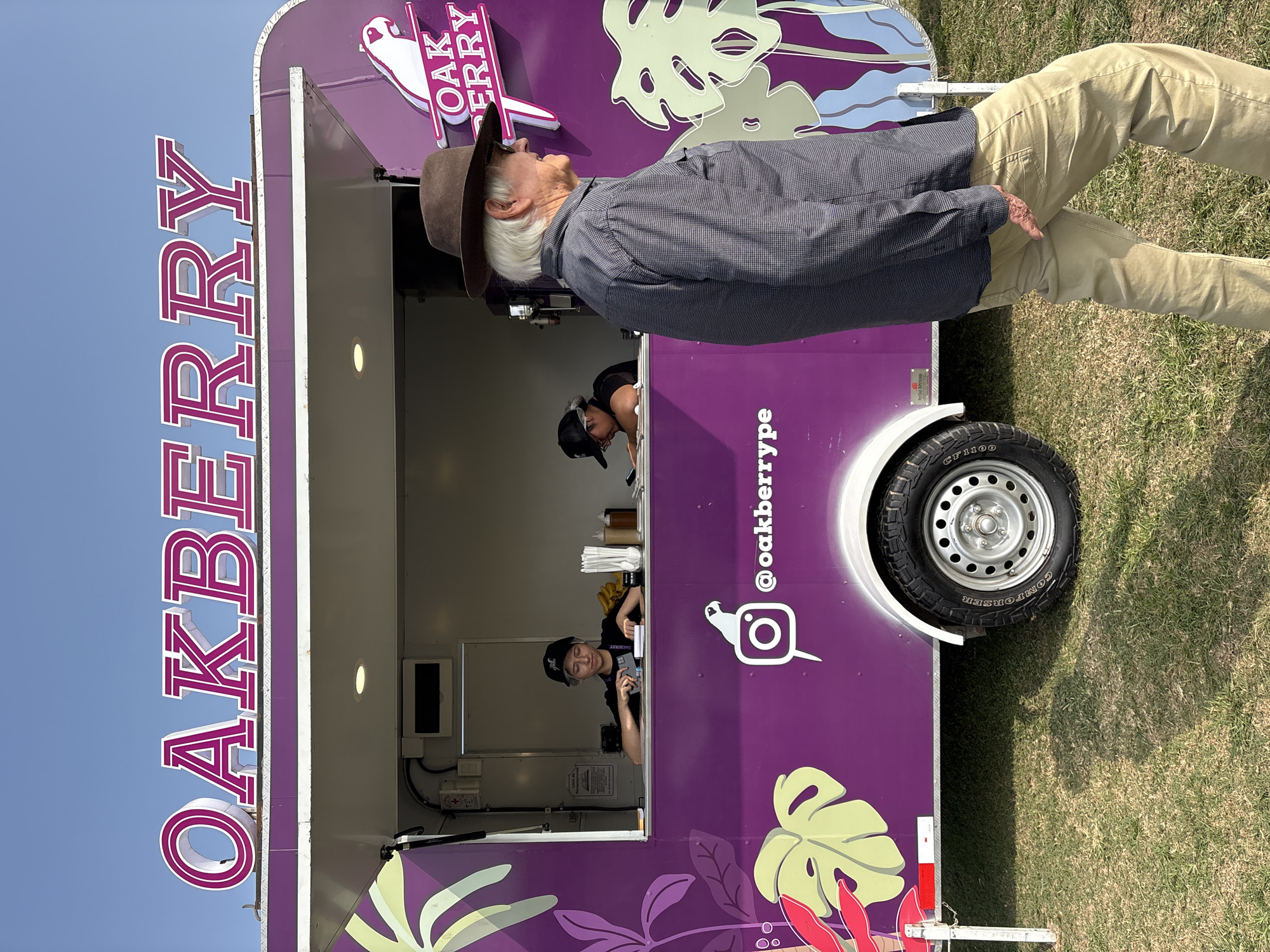 The image depicts a bright, sunny day in Lurín, Peru, with a vibrant purple food truck named "OAKBERRY" as the central focus. The truck features large, stylized text spelling out its name in pink and white. A logo of a white bird on a pink circle, along with the Instagram handle "@oakberrytype", is visible on the side of the truck. Tropical leaf designs in shades of green and pink adorn the truck's exterior.
Two employees are visible inside the truck's service window. One, wearing a black baseball cap, is focused on something on the counter, possibly taking an order or preparing food. The other employee, also wearing a black cap, is partially obscured but appears to be working behind the counter.
In the foreground, an elderly man with white hair and a brown hat is walking past the food truck, his back mostly to the camera. He is wearing a dark, checkered long-sleeved shirt and light brown pants.
The food truck is parked on a grassy area, suggesting an outdoor event or location. The sky is clear and blue. The overall scene is casual and appears to be a typical day at a food vendor, possibly at a market or festival in Lurín.