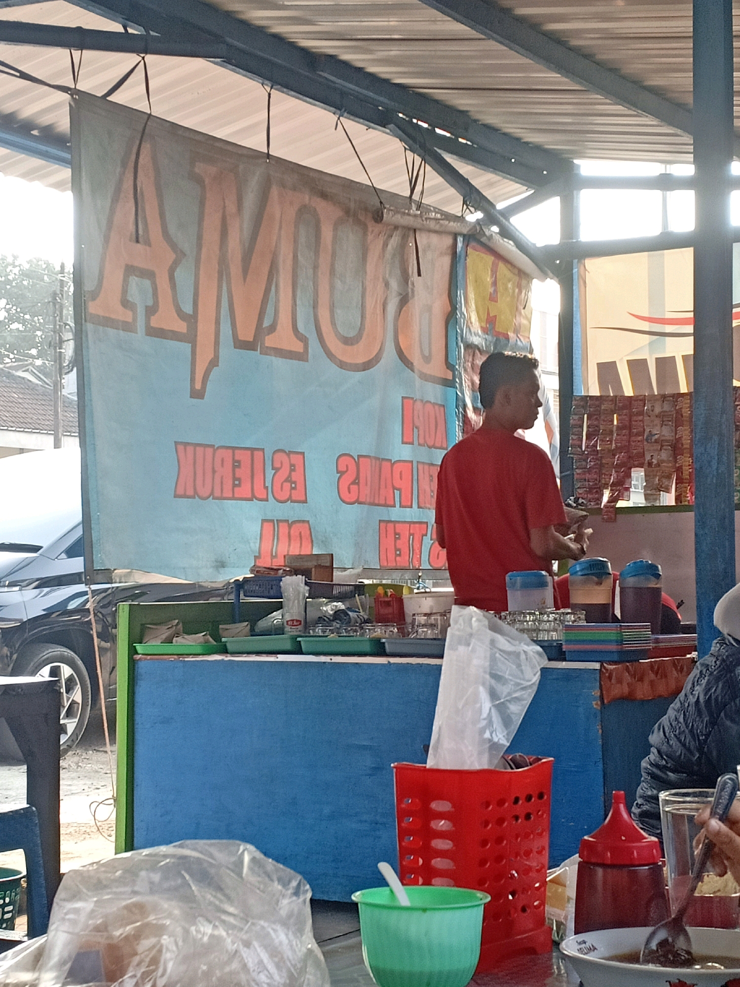 The image depicts an outdoor food stall in Jatisrono, Indonesia. A man in a red t-shirt is standing behind a blue counter, appearing to serve customers. In front of him are various items including drinks in dispensers, stacks of bowls, and plastic bags. A large banner with orange lettering on a light blue background hangs behind the stall, displaying Indonesian text, likely advertising the food or drinks sold. To the left, a black car is partially visible. The environment appears to be a roadside setting under a corrugated metal roof, with sunlight filtering through. The overall scene suggests a typical street food vendor operation.