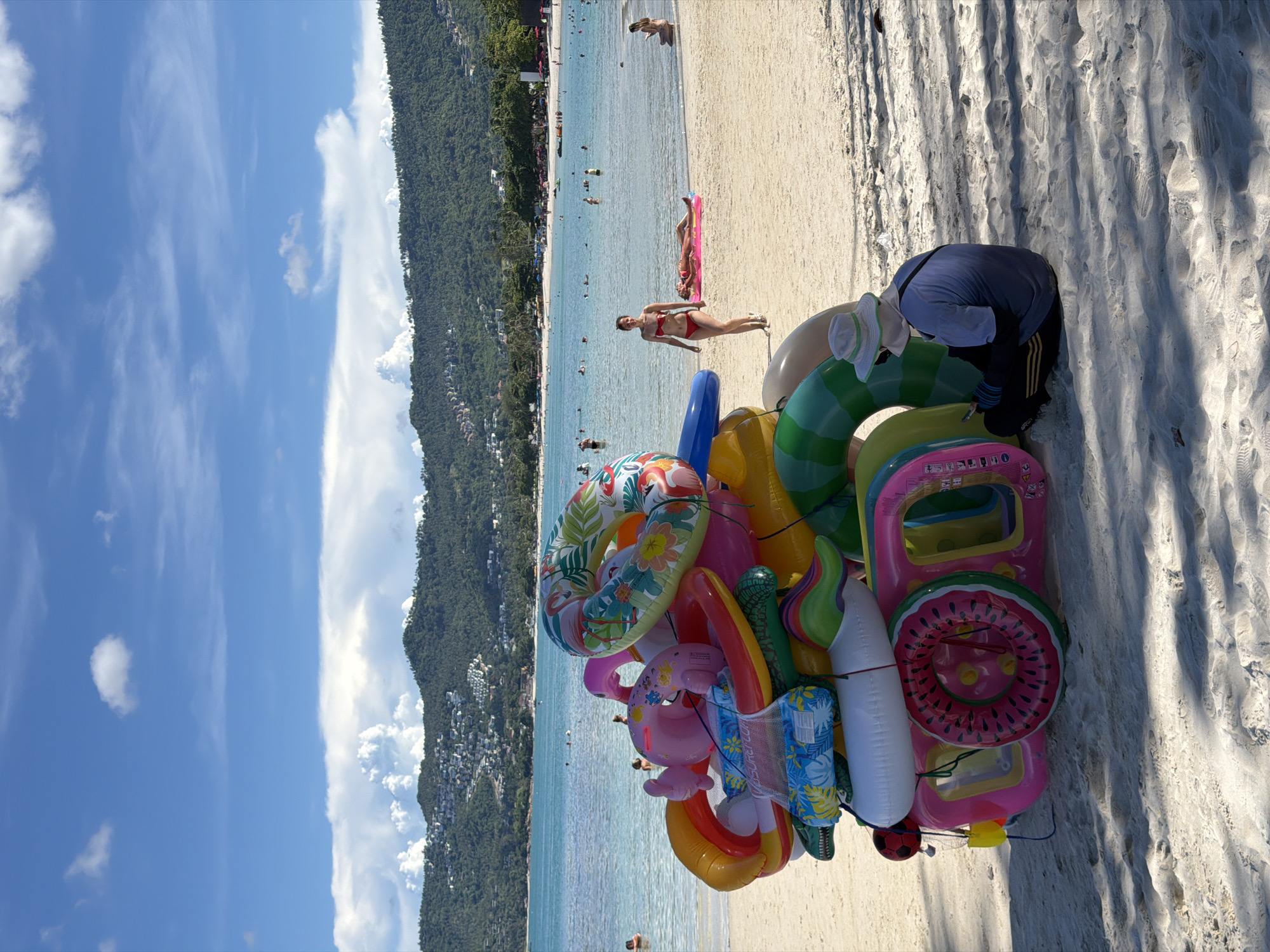 The image depicts a sunny day on Baan Chaweng beach in Thailand. In the foreground, a large, colorful display of inflatable pool toys, including donuts, flamingos, and novelty shapes, is piled high on the sand. To the right, a person wearing a light-colored hat and a blue shirt sits on the sand, looking at their phone. In the mid-ground, the turquoise water of the ocean is visible, with several people swimming and relaxing. A woman in a red bikini walks along the shore, and another person is lying on a pink inflatable raft. The background shows a lush, green, mountainous landscape dotted with buildings, extending to the horizon under a bright blue sky with scattered white clouds.