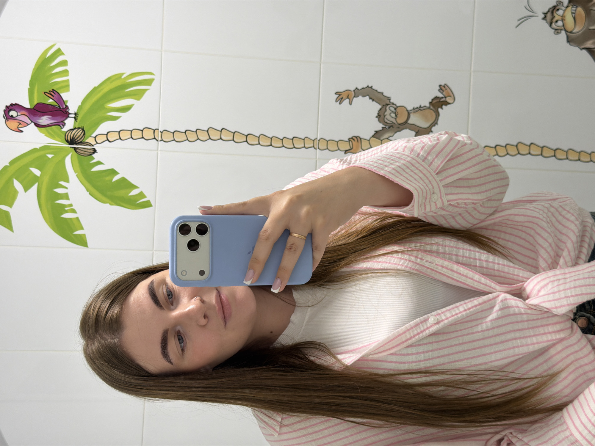 A woman is taking a selfie in what appears to be a bathroom. She is holding a blue smartphone in her right hand. She has light brown hair, fair skin, and is wearing a white top with a pink and white striped shirt or jacket on top. Her nails are manicured with a French tip. The background is a white tiled wall with cartoonish drawings of palm trees, a bird, and monkeys on a vine. It is assumed the location is in the city of Chaikovsky, Russia.