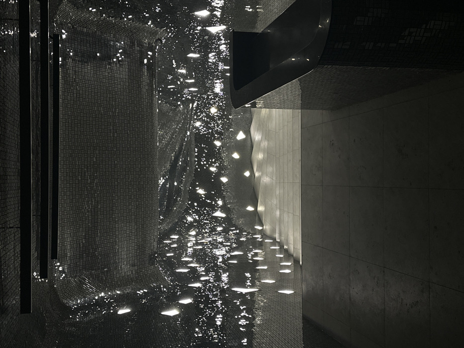 The image depicts an interior view of a modern architectural space, identified as Zaryadye Park in Moscow, Russia. The environment is characterized by an absence of people and a distinct design. The walls and a portion of the vaulted ceiling are covered entirely in small, dark, reflective tiles, creating a shimmering, textured surface that reflects numerous small, bright, irregularly shaped points of light. The left wall and parts of the ceiling exhibit significant, wave-like undulations. The floor consists of large, light-grey square tiles. In the foreground on the right, a dark, curved counter or bar structure, also surfaced with small reflective tiles, extends into the space. Its top surface is a smooth, dark, glossy plane. Towards the rear, a light-colored staircase ascends with several steps, illuminated by overhead light, integrating with the floor's material. Overhead, two dark, linear fixtures are visible against the tiled ceiling. The overall lighting is dim, emphasizing the reflections and architectural forms.