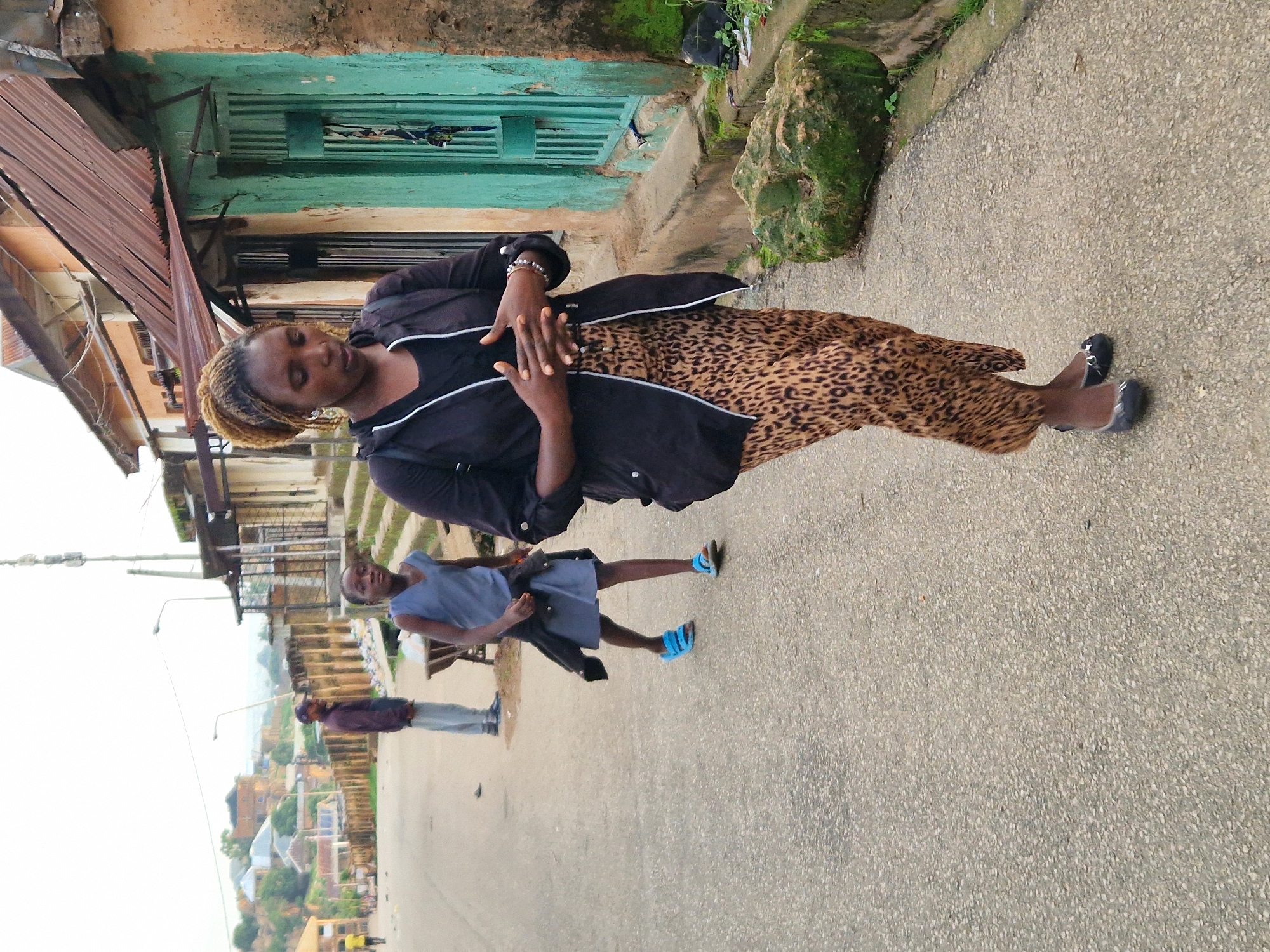 The image depicts an outdoor scene on a paved street in an urban environment, likely in Africa based on the architecture and attire. In the foreground, a woman with braided hair is walking towards the viewer, her hands clasped in front of her. She is wearing a black jacket over a black top and a leopard-print skirt. She is also wearing dark, pointed ballet flats. To her left and slightly behind her, a young girl is walking away from the viewer. The girl is wearing a blue sleeveless dress over a grey skirt and blue sandals. Behind the girl and to the left, a man is standing in the street, facing away from the viewer. He is wearing a dark jacket, light grey pants, and a baseball cap. The background shows a street lined with buildings that have corrugated metal roofs and painted facades, some appearing aged. There are also power lines and utility poles visible. The ground is a mix of paved asphalt and concrete sidewalks. The overall impression is a scene of everyday life in a developing urban area.
