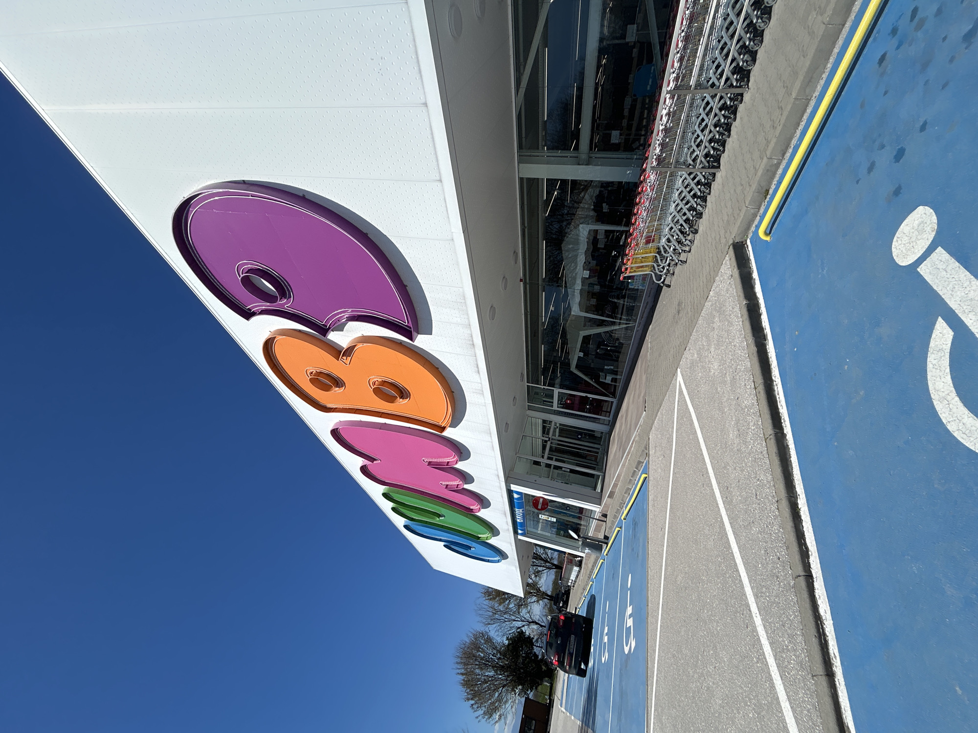 The image shows the exterior of a store, likely in Plovdiv, Bulgaria, based on the signage. The store has a large, colorful logo, with the letters "CBA" stacked vertically in purple, orange, pink, green, and blue. To the right of the store is a parking area painted blue, marked with a wheelchair symbol. Shopping carts are aligned on the edge of the parking area. A black car is parked in one of the handicap parking spots. A tree is visible in the background. The sky is clear and blue.