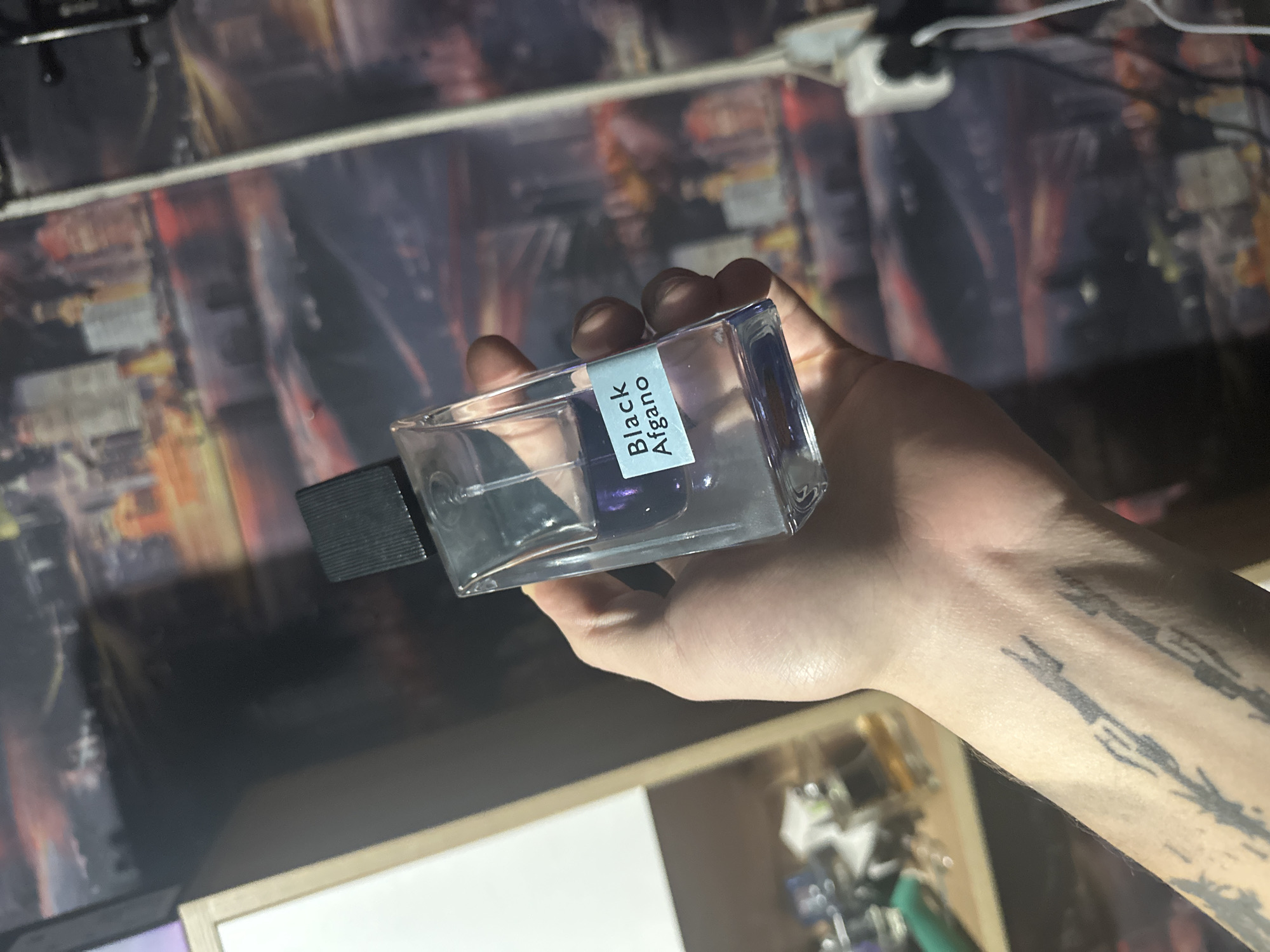 A person's left hand holds a partially full bottle of Nasomatto Black Afgano perfume. The bottle is clear glass with a black cap and a light blue label. The hand has a tattoo on the wrist and forearm. The background is blurred, showing a desk with a cityscape design on the top surface, a white drawer unit, and some electrical cords and devices. This image does not provide any information to identify the location as Gorno-Altaysk, Russia.