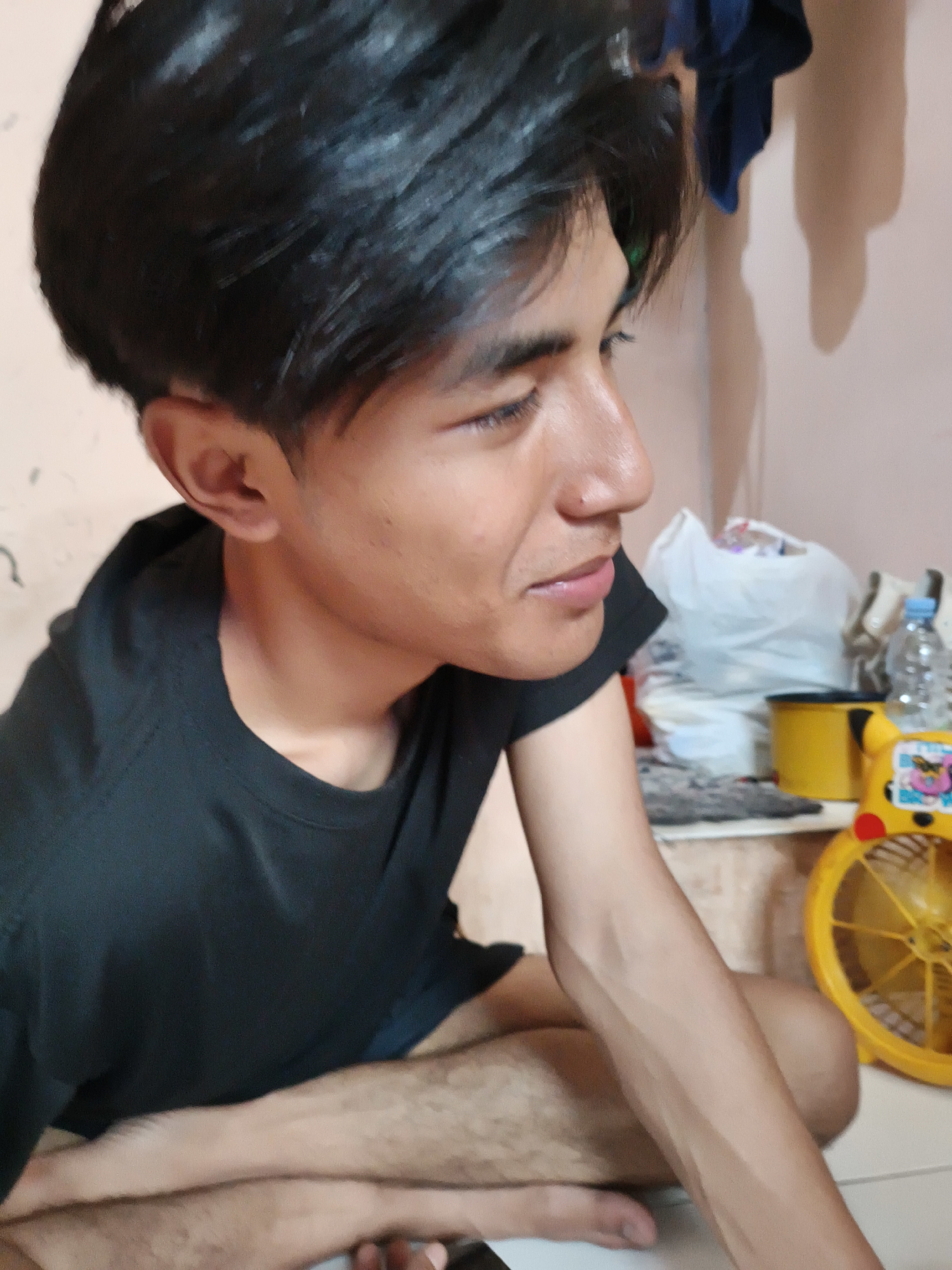 The image shows an individual with dark hair and a black t-shirt sitting cross-legged on the floor. The person is facing to the right, with their arm resting on their leg. In the background, there is a cluttered surface with a yellow container, a water bottle, a white plastic bag, and a small yellow fan with a Pikachu design. The wall is a light pink color. The location is plausibly in Cianjur, Indonesia given the facial features of the person. No other specific details confirm this location.