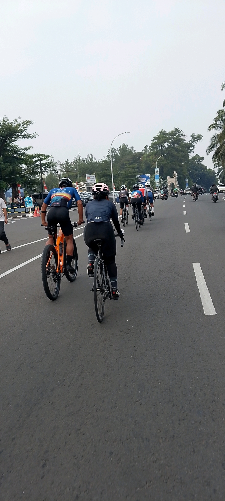 The image shows a group of cyclists on a road in Gunung Putri, Indonesia. In the foreground, two cyclists are riding side-by-side. The cyclist on the left is wearing a blue and orange cycling jersey and black shorts, and is riding an orange bicycle. The cyclist on the right is wearing a dark blue cycling jersey and black leggings, and is riding a black bicycle. Both cyclists are wearing helmets. In the background, a procession of cyclists and motorcyclists are visible. The road is paved and has white lines marking the lanes. Trees and buildings line the side of the road. The sky is overcast.