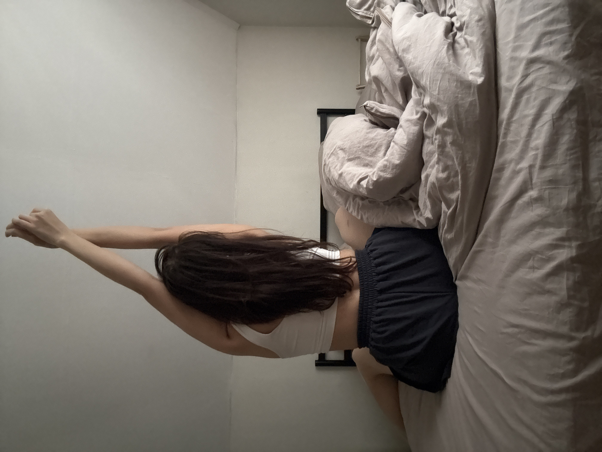 A woman lies on a bed in a room. She is wearing a white top and blue shorts. Her arms are stretched overhead. The bed has grey sheets and is positioned against a white wall. There is a dark, rectangular object (possibly a headboard or picture frame) near the head of the bed. The location is Chaikovsky, Russia, but the image does not provide conclusive forensic data to confirm this claim.