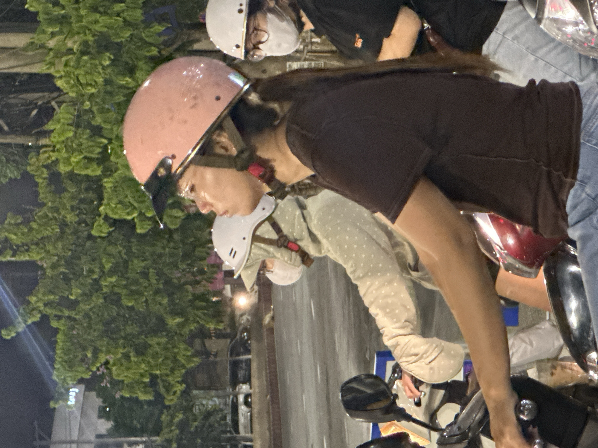 The image shows three individuals on motorbikes at night, likely in a city environment. The closest person, on the right, is wearing a dark t-shirt and blue jeans, with a pink helmet. They are looking upwards and to the left. Behind them, another person is wearing a light-colored, hooded garment with white polka dots and a white helmet. This person is also looking forward or slightly to the left and appears to be wearing a face mask. Further in the background, a third person is visible wearing a white helmet and glasses, also with a face mask and a dark shirt. The background features trees, streetlights, and what appear to be buildings or other structures. The presence of motorbikes and the attire of the individuals suggest a common mode of transportation in Vietnam. The road surface is visible, indicating an outdoor setting. Without specific landmarks, pinpointing the exact location within Đà Nẵng is not possible from the image alone.