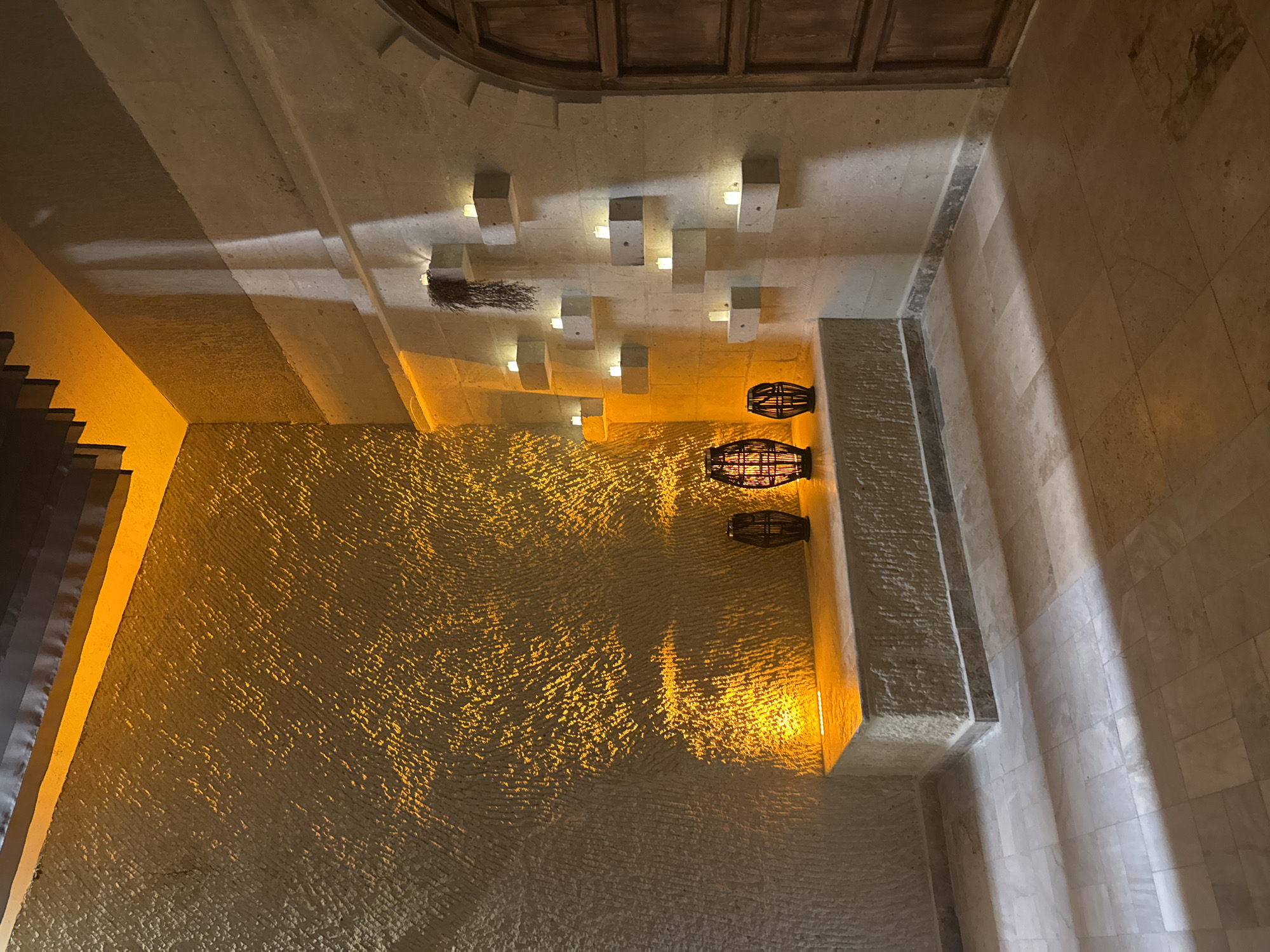 The image shows an interior architectural space with textured walls lit by warm, artificial lighting. The ceiling has a series of square, blocky light fixtures and three hanging, cage-like decorative elements. The space appears to be in Ürgüp, Turkey, given the style of architecture and lighting. No people are visible.