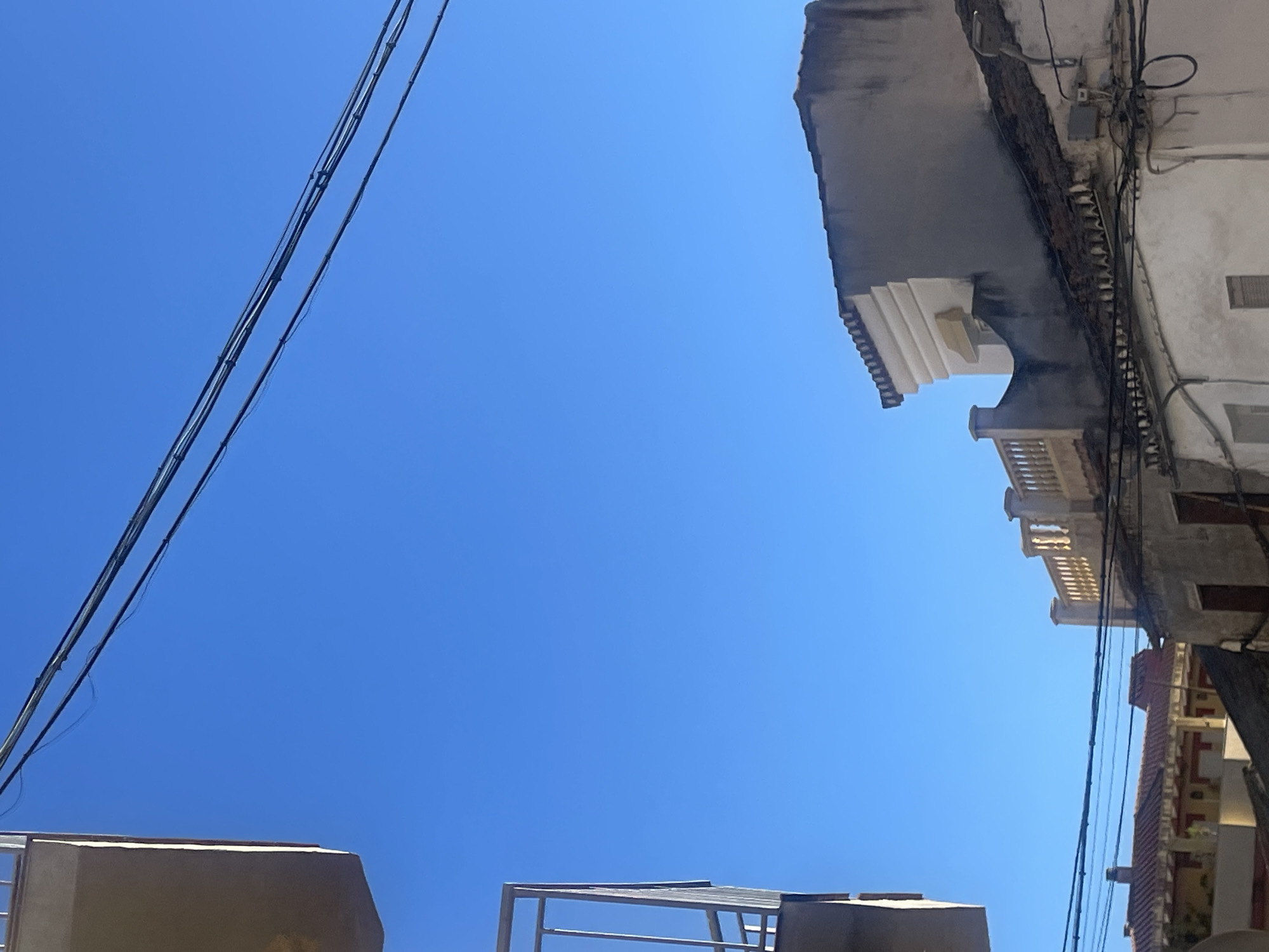 The image captures a view of buildings and utility lines against a clear blue sky in Atarfe, Spain. In the foreground, a sliver of a light-colored structure, possibly a balcony, is visible on the left. Overhead, several dark electrical wires are strung across the frame, with one set of wires running diagonally from the upper left to the upper right. Below the wires, the rooftops of several buildings are visible. On the right side, a building with a light-colored decorative facade and a balustrade is prominent. Further to the left, the tiled roof of another building with red and yellow accents can be seen. The overall scene depicts an urban environment, likely residential, with clear skies and an emphasis on architectural details and overhead infrastructure. There are no people or animals visible in the image.