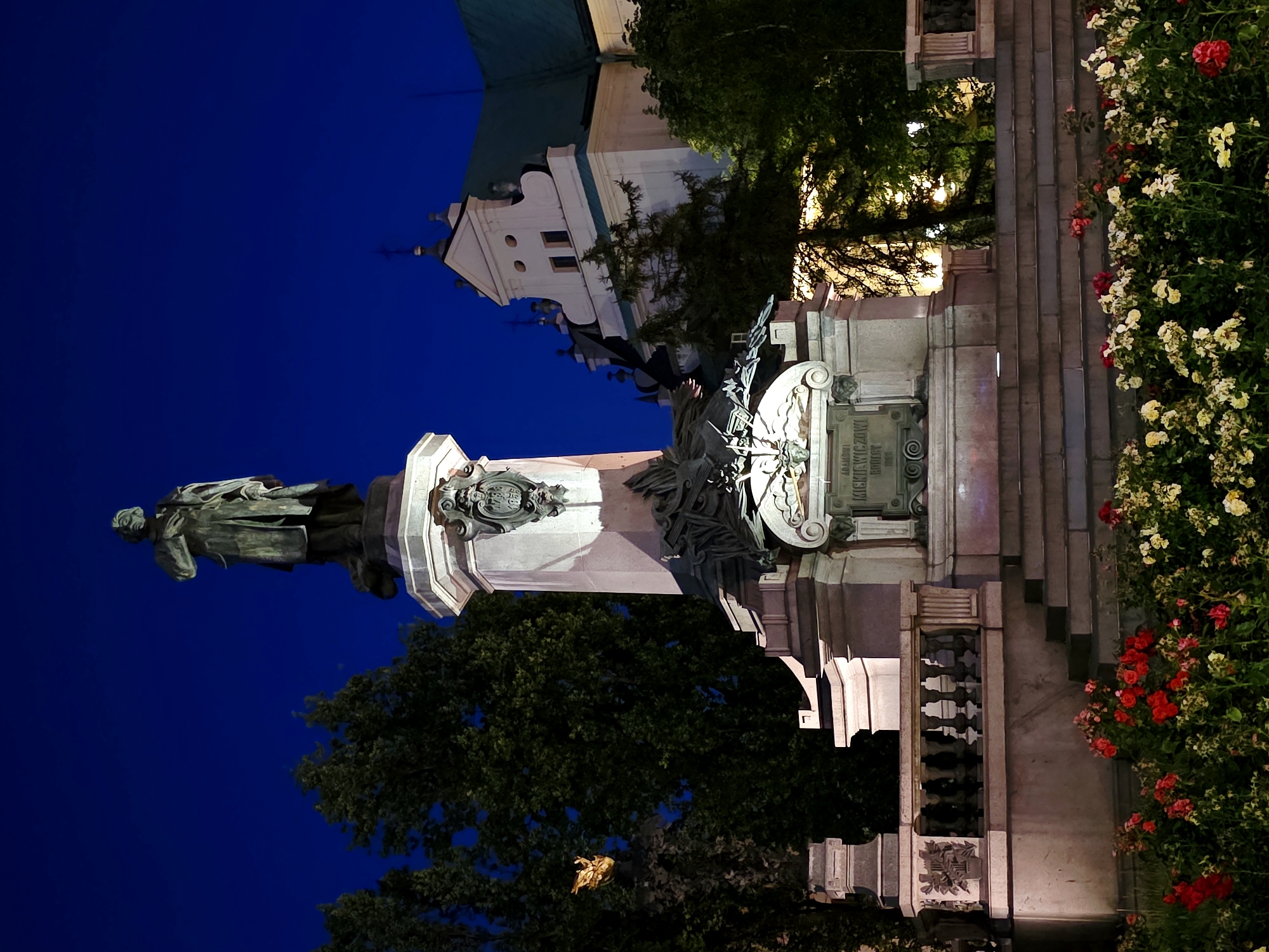 This is a full-shot, eye-level, outdoor, nighttime photograph of the Adam Mickiewicz Monument in Warsaw, Poland. The monument, made of bronze and stone, depicts a standing male figure on a large, multi-tiered pedestal. The pedestal has ornate carvings and inscriptions. In the foreground, there is a bed of red and white roses. Behind the monument, there are trees and a building with a green roof. The sky is a deep blue. The lighting suggests artificial illumination. The monument is a prominent historical landmark in the city.