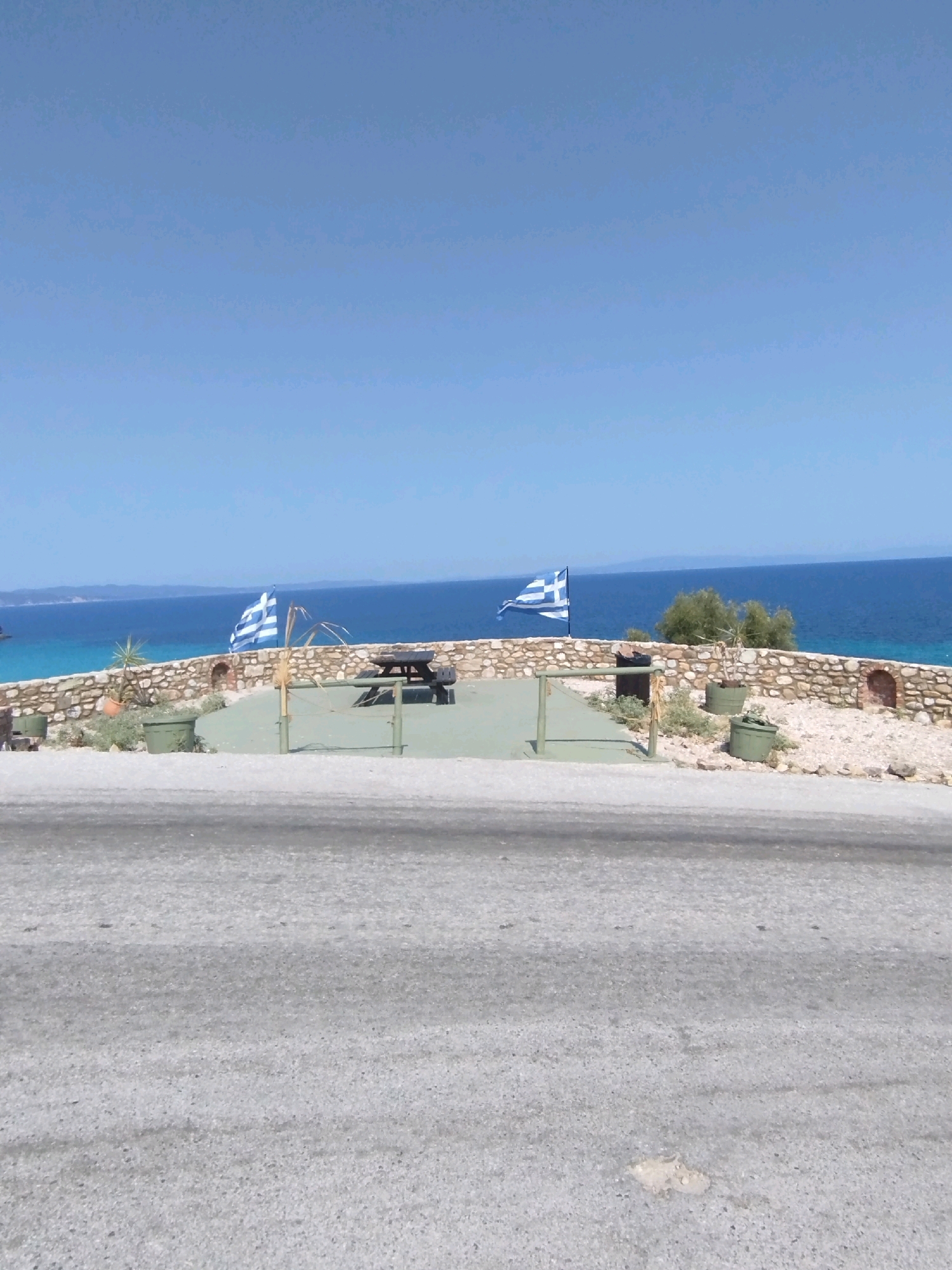 The image depicts a view of the sea and a paved area with a stone wall, two Greek flags, a picnic table, and several green planters. The scene appears to be a scenic overlook or a rest stop. The sea is a vibrant blue, with a distant shoreline visible. The sky is clear and bright blue. The foreground shows a grey asphalt road. The location is identified as being in the Sithonia Municipal Unit, Greece. There are no people visible in the image.