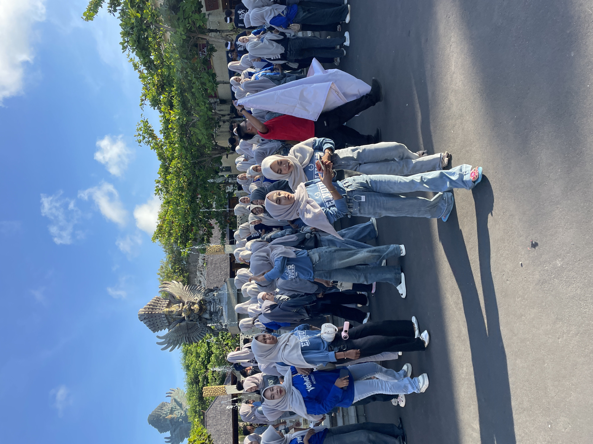 A crowd of people are lined up in Kuta Selatan, Indonesia. Most of the people are wearing a light-colored hijab and a light blue or dark blue sweatshirt, blue jeans, and sneakers. In the background is a large statue of an eagle and a lush green area. Some people are holding a large white cloth.