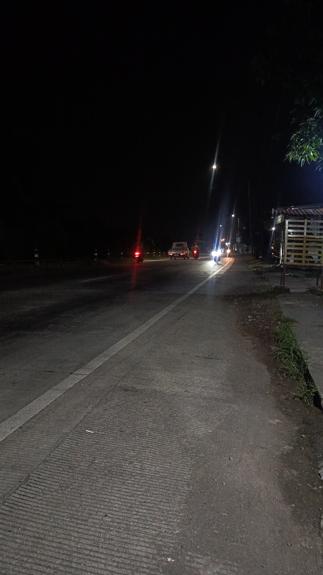 The image depicts a night scene on a road in Kajeksan, Indonesia. The foreground shows a paved road with textured tire grip sections and a white painted lane marker. To the right is a narrow sidewalk with some grass growing along the edge.
In the background, there are multiple vehicles on the road. From left to right, there is a motorcycle with red taillights, a small flatbed truck, and another motorcycle, also with red taillights. Further down the road, there are two more motorcycles with headlights on.
Streetlights illuminate the scene. A small, wooden structure sits on the right side of the road. The sky is dark. The overall lighting is dim, and visibility is limited.
