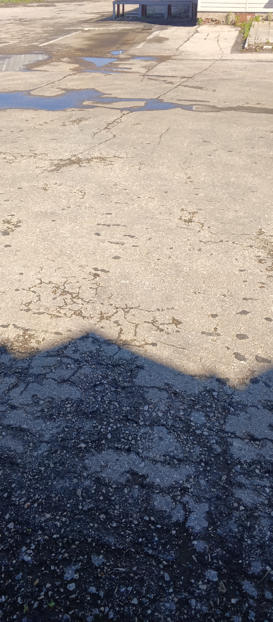 The image shows an outdoor scene, likely a cracked asphalt parking lot, with puddles of water and a large shadow cast across the ground. In the background, there is a building with a white facade and what appears to be a covered structure with support beams. The ground is a mix of light gray and darker gray asphalt, with visible cracks and gravel. The shadow, which appears to be from a building or structure, creates a distinct line dividing the sunlit and shadowed areas of the asphalt. There are no people or animals visible in the image. The environment appears to be overcast or experiencing partial shade, possibly during the day.