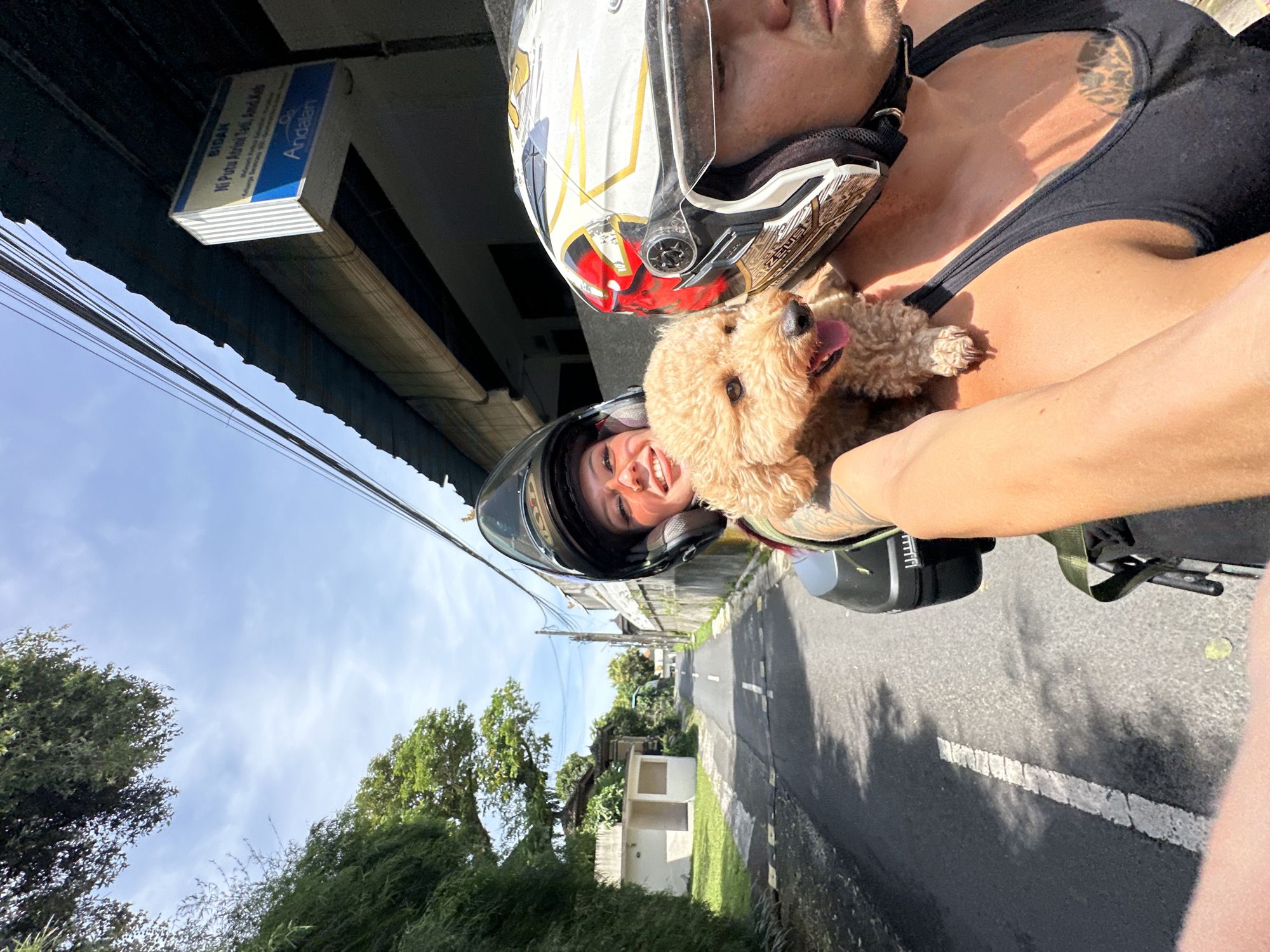 A medium shot depicts two individuals and a dog riding a motorcycle in North Kuta, Indonesia. The person in the foreground wears a black tank top, a helmet, and holds a tan-colored dog. The second person, behind them, wears a helmet and smiles. The motorcycle travels down a street with trees and buildings on either side. A sign reading "Bidari Ni Pita Atria Tati, Amd Keb" and "Andalan" is visible. Power lines run above the street.