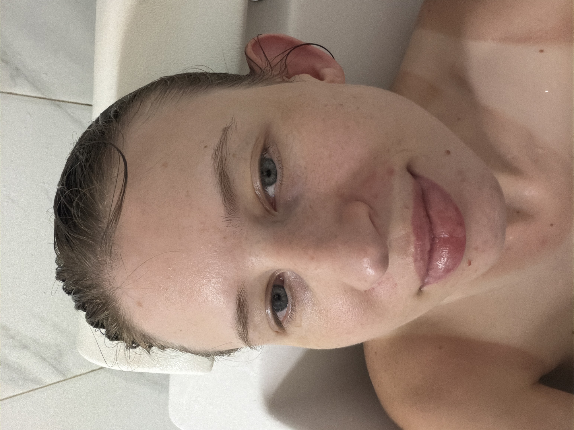 A close-up, head-on shot captures a woman's face and upper body from a slightly elevated perspective. Her hair, dark and wet, is slicked back, revealing a fair complexion dotted with freckles. Her eyes, a light blue-grey, are open and looking directly at the camera. Her lips are slightly parted, showing a glossy, pinkish hue. The background consists of white tiled walls and what appears to be a white bath cushion or headrest. The lighting is bright, highlighting the water droplets on her skin and the sheen on her lips.