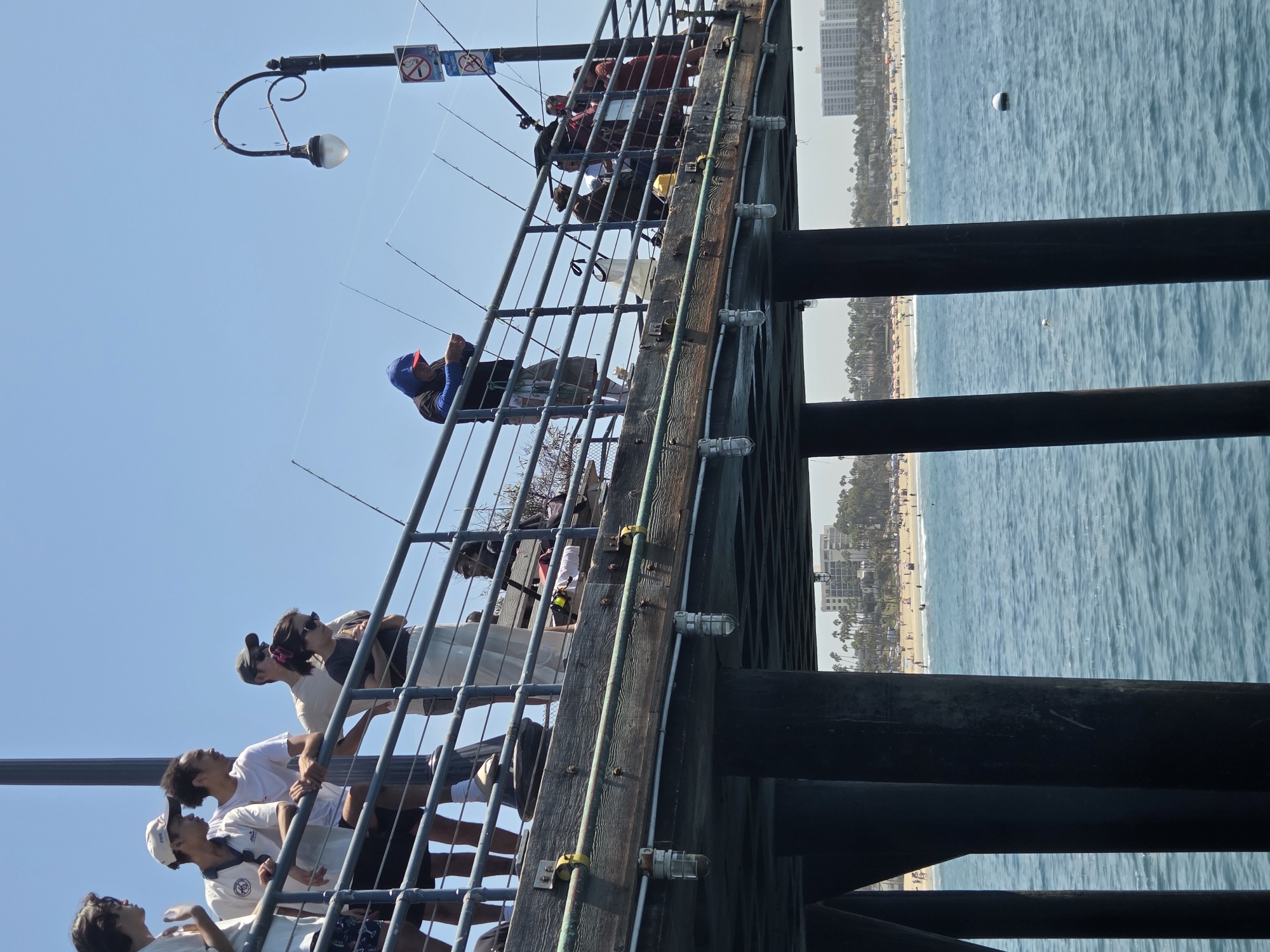 The image shows a view from the underside of a pier in Santa Monica, California, looking out towards the ocean and beach. Several people are visible on the pier, most of whom appear to be fishing. The ocean is a clear blue, with gentle waves. The beach in the background is crowded with sunbathers and umbrellas. The sky is clear and blue, indicating a sunny day. The pier itself is made of weathered wood and supported by dark, thick pilings. There are several fishing rods set up along the railing of the pier.