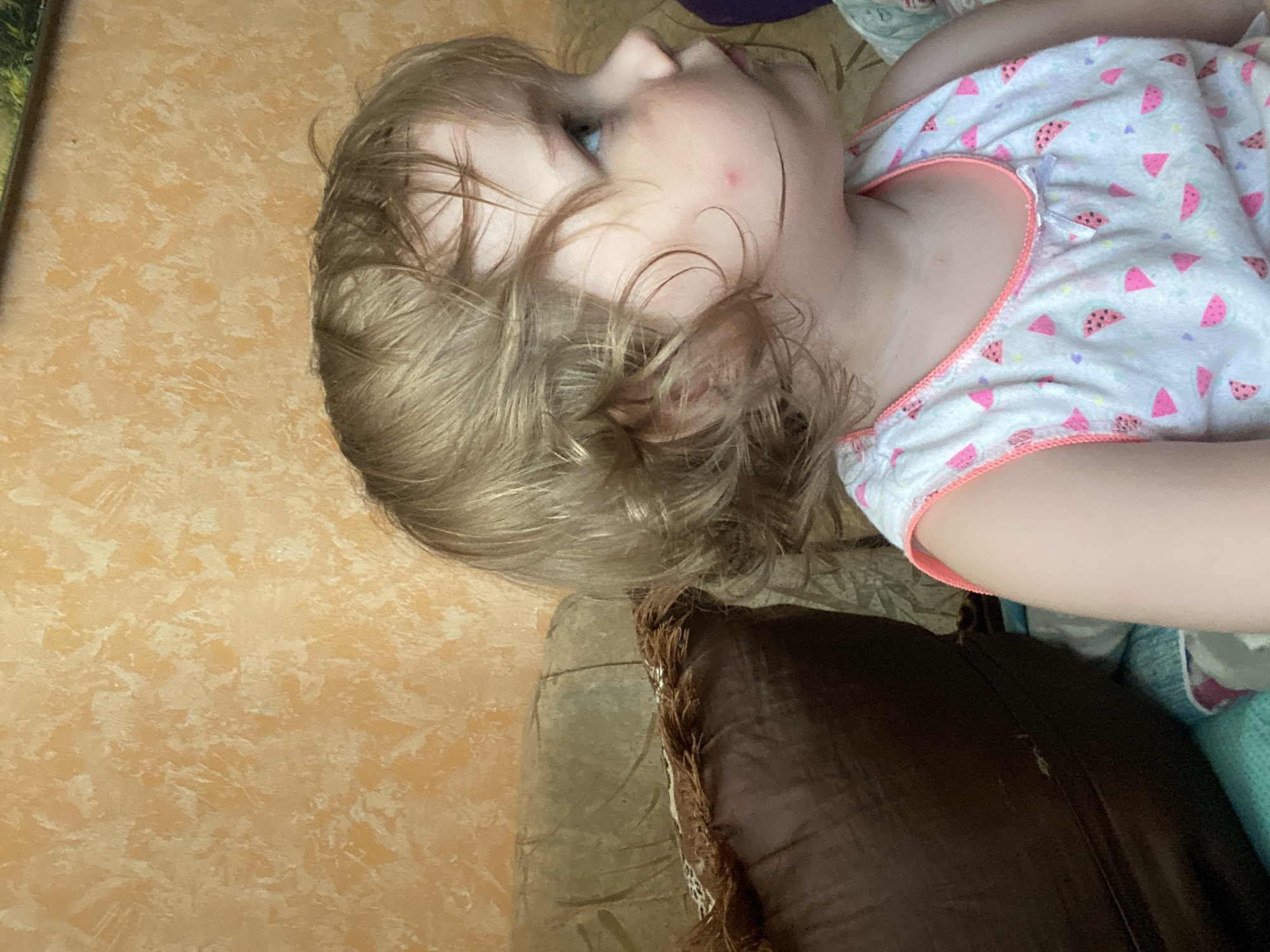 A young blonde-haired child is seated on a couch, facing right. The child is wearing a white sleeveless shirt with a pink watermelon pattern and a pink trim. The child's hair is curly and slightly messy. The background shows a textured orange wall and a portion of a brown couch with a dark brown cushion. The lighting is warm and appears to be natural or from indoor sources.
