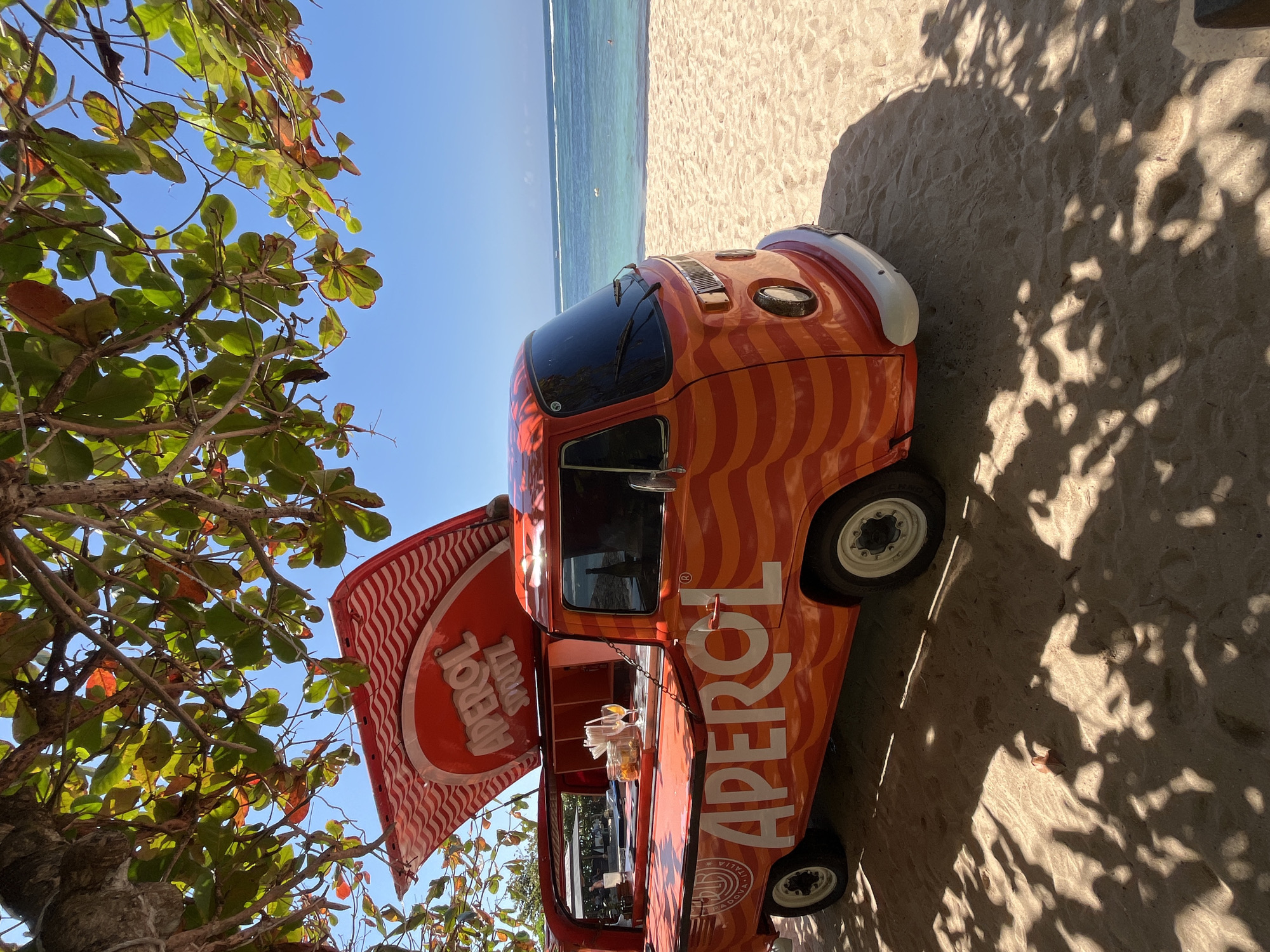 An orange Volkswagen Type 2 van, modified into a mobile bar with "APEROL SPRITZ" branding, is parked on a sandy beach in Kuta Selatan, Indonesia. The van features wavy orange stripes and a raised red and white awning. Two glasses with orange drinks and a jar of what appears to be oranges are visible on the bar counter. The ocean, with gentle waves and a clear blue sky, stretches out in the background. Tree branches with green and red leaves hang over the scene, casting dappled shadows on the sand. There are no people clearly visible in the immediate vicinity of the van.