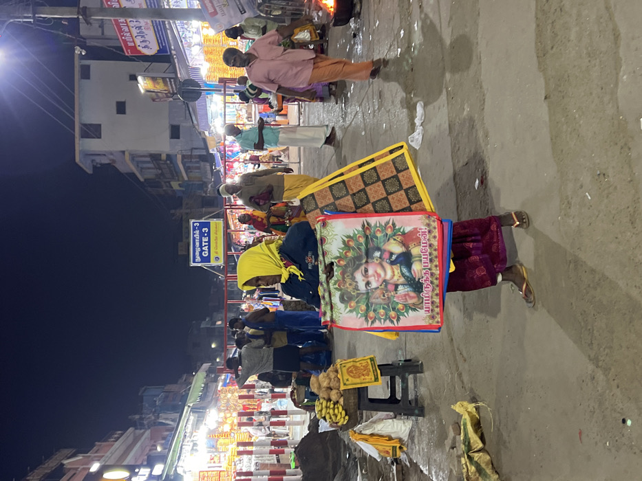 This nighttime image captures a lively street scene in Palani, India, bustling with people and market stalls. The dark sky is illuminated by numerous artificial lights from shops and a prominent street light on the left.
In the foreground, a person wearing a yellow headscarf and a dark top is holding several colorful reusable bags. One bag prominently displays a vibrant image of Lord Murugan (Kartikeya) with a peacock, a significant deity in South Indian Hinduism. Tamil text on this bag includes a devotional phrase like "யா முருகையா" (Ya Murugaiya). Another bag features a brown and black geometric pattern. Near this person, a small stall offers bananas and coconuts, common items in temple towns.
Further back, several other people are visible. A man in a pink shirt and orange dhoti stands near a small fire, possibly for an offering or ritual. Other individuals are walking or standing amidst numerous brightly lit market stalls lining the street, suggesting an active commercial area. Red and white striped barricades are present in the mid-ground, likely used for crowd control or defining a pathway.
A blue sign, clearly visible, features text in both Tamil ("நமச்சிவாயன் - 3") and English ("GATE - 3"), indicating "Gate 3" of a complex, possibly a temple, and also reads "© Swamimalai Photos". The scene strongly suggests the vicinity of a major pilgrimage site, typical of Palani, where devotees gather and shop for religious items and offerings. The atmosphere is busy and illuminated against the night.