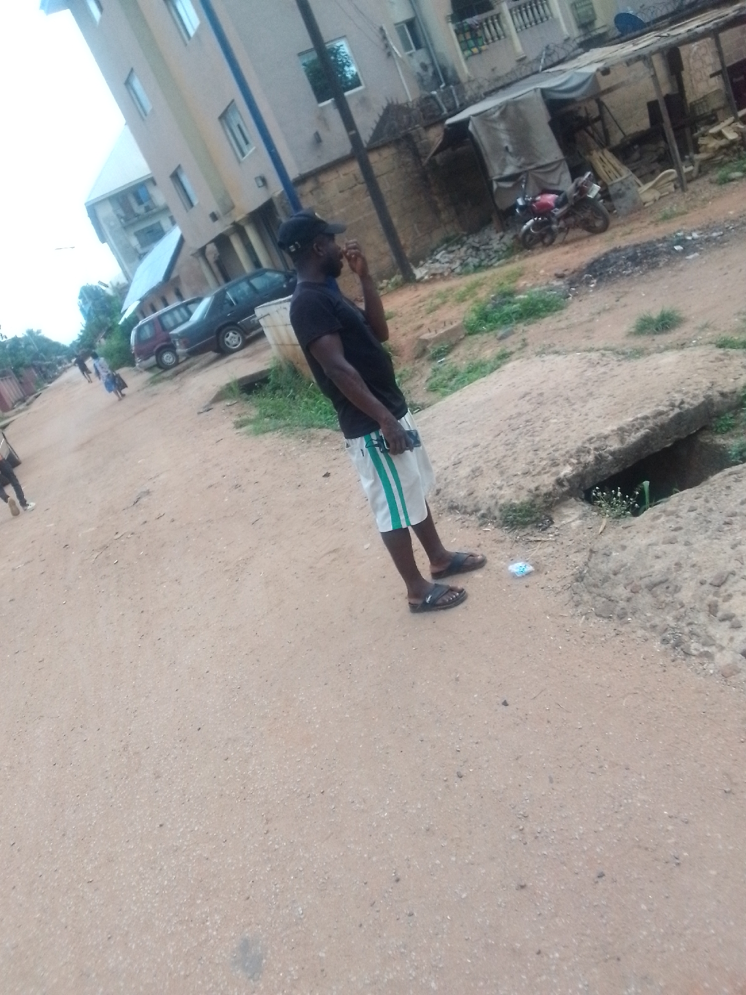 The image shows a man standing on a dirt road in an urban environment. He is wearing a black shirt, white shorts with green stripes, a dark cap, and sandals. He appears to be talking on a cell phone. The road is unpaved and shows signs of wear and tear. Adjacent to the man, a concrete structure covers a drainage ditch. The surrounding area includes buildings, parked vehicles, and a motorcycle parked near what appears to be a makeshift shelter. Other individuals are visible in the distance on the road.