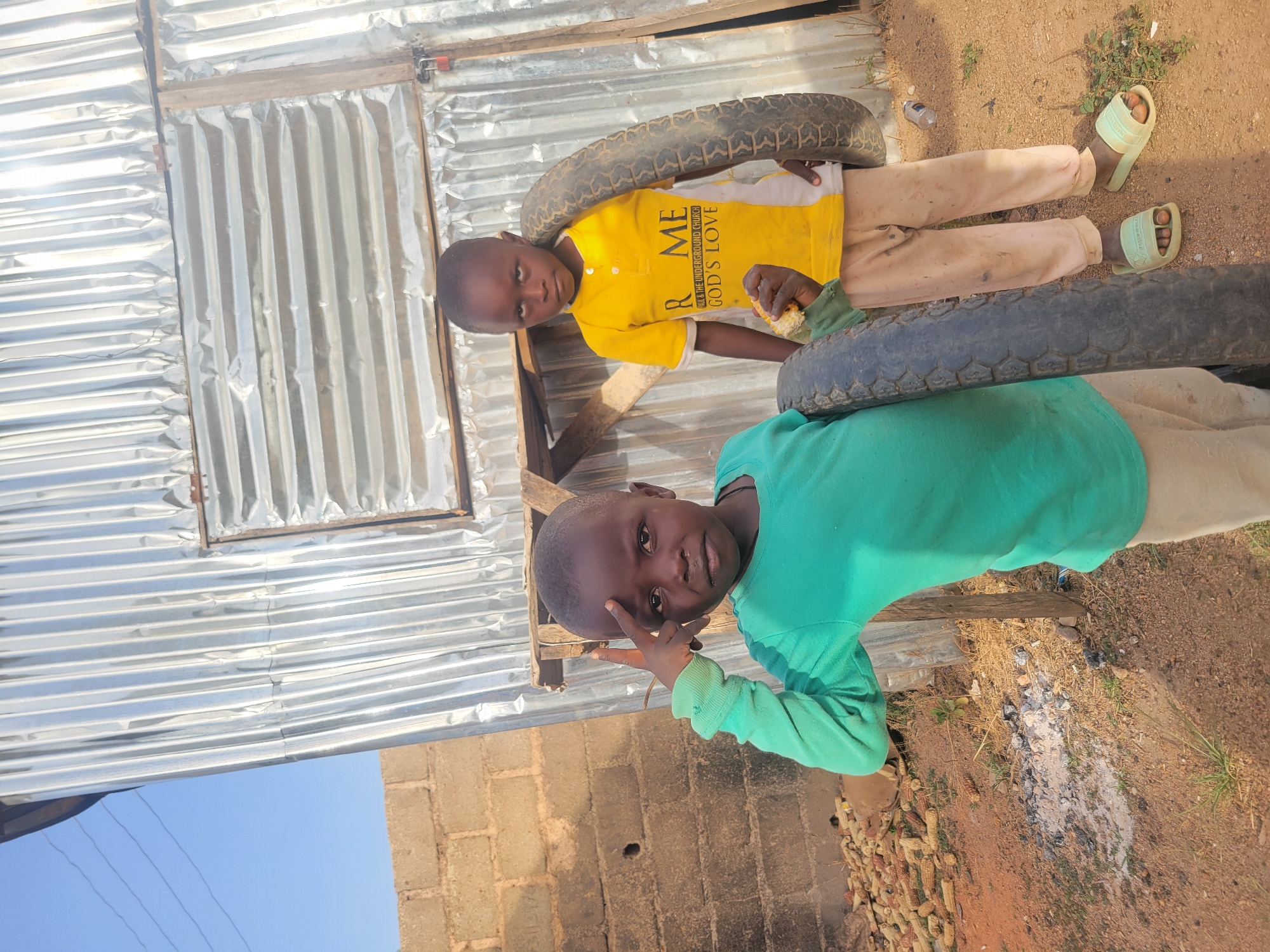 Two young boys are depicted outdoors in Barakin Lerawi, Nigeria, under bright daylight. The boy on the left wears a yellow and white polo shirt with the visible text "ME R & THE UNDERGROUND CHURCH GOD'S LOVE," light brown trousers, and light green slides. He has a tire draped over his shoulder and is holding a piece of corn on the cob. He looks towards the camera with a neutral expression.
The boy on the right wears a green long-sleeved shirt and has a tire around his neck/shoulder. He makes a peace sign with his right hand and looks towards the camera with a subtle smile.
They are standing on a dirt ground with some sparse vegetation and scattered small rocks. In the background, there is a structure constructed from corrugated metal sheets and wooden beams, suggesting a simple building or shelter. A section of a concrete block wall is also visible. Near the bottom right, there is a pile of dried corn cobs, and a discarded plastic water bottle is on the ground. A clear blue sky is visible at the upper left, indicating fair weather. The scene reflects a casual moment in a rural or semi-urban outdoor environment.