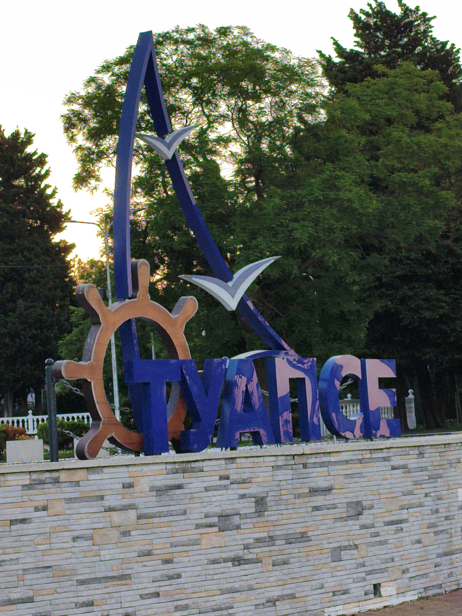 The image shows a large, blue, three-dimensional sign that reads "TUAPSE" in Russian Cyrillic letters. The sign is decorated with elements suggestive of a seaside or port town, including a ship's wheel and two stylized white birds, likely representing seagulls, in flight. The letters themselves have a textured, patterned design in shades of purple and pink.
The sign is mounted on a low, multi-layered stone wall, constructed from rectangular blocks of varying shades of gray, beige, and tan. Behind the sign, lush green trees form a dense backdrop, with the tops of several tall evergreen trees visible on the left and right edges of the frame. A bright sky, appearing overcast or at dusk, is visible in the upper left portion of the image. Sunlight can be seen filtering through the trees on the left, casting a warm glow.
In the background, beyond the sign and the stone wall, a white ornate fence can be seen, suggesting a park or public space. A lamppost is also visible behind the sign. The overall scene evokes a welcoming atmosphere, characteristic of a tourist destination. The time of day appears to be late afternoon or early evening, based on the soft lighting and the hint of sunset colors through the trees. The weather seems calm and pleasant, with no indication of harsh conditions. The presence of the ship's wheel and seagulls strongly suggests a coastal location, consistent with Tuapse being a port city on the Black Sea. No people are visible in the frame.
