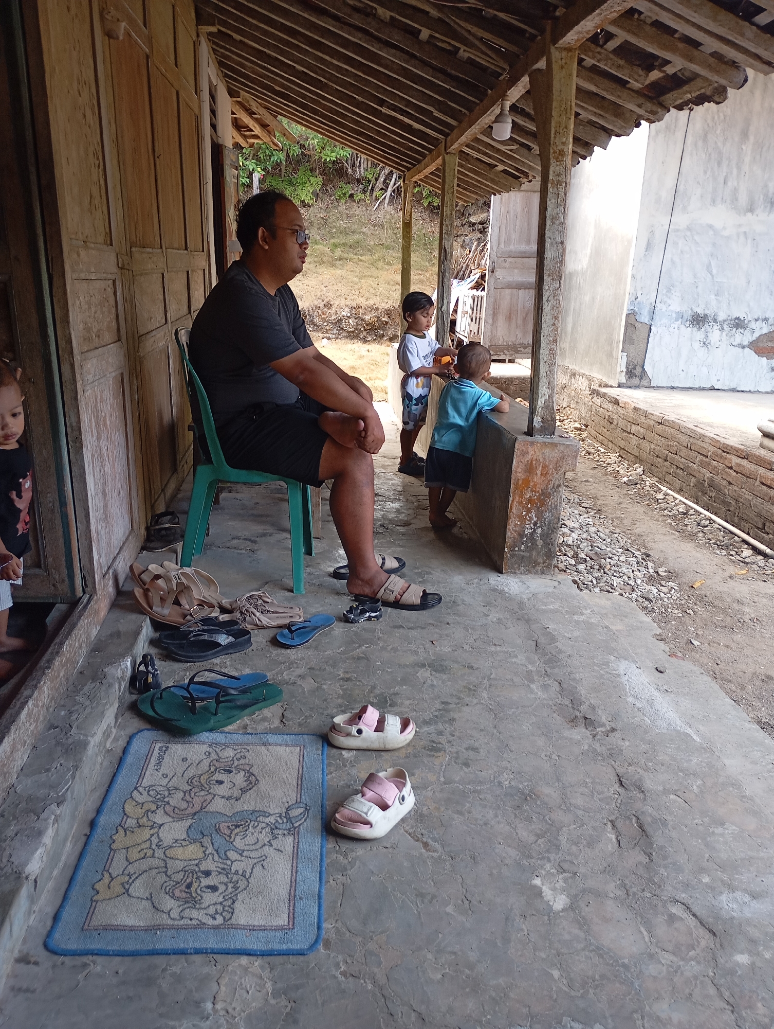 In Ketepung, Indonesia, a man wearing glasses, a dark t-shirt, and shorts sits on a green plastic chair in front of a wooden structure. In front of him, several pairs of sandals are scattered on the concrete floor, including brown and black flip-flops, and a pair of blue flip-flops. To his right, two young children stand near a concrete pillar; one child wears a white t-shirt with a graphic and patterned shorts, and the other wears a blue t-shirt and dark shorts. A light-colored wall stands to the right of the children, with a brick foundation. A blue mat with Disney characters is placed on the concrete in front of the man. In the background, a grassy area with trees is visible, suggesting a rural or semi-rural setting. The overall scene depicts a casual outdoor gathering on a sunny day.