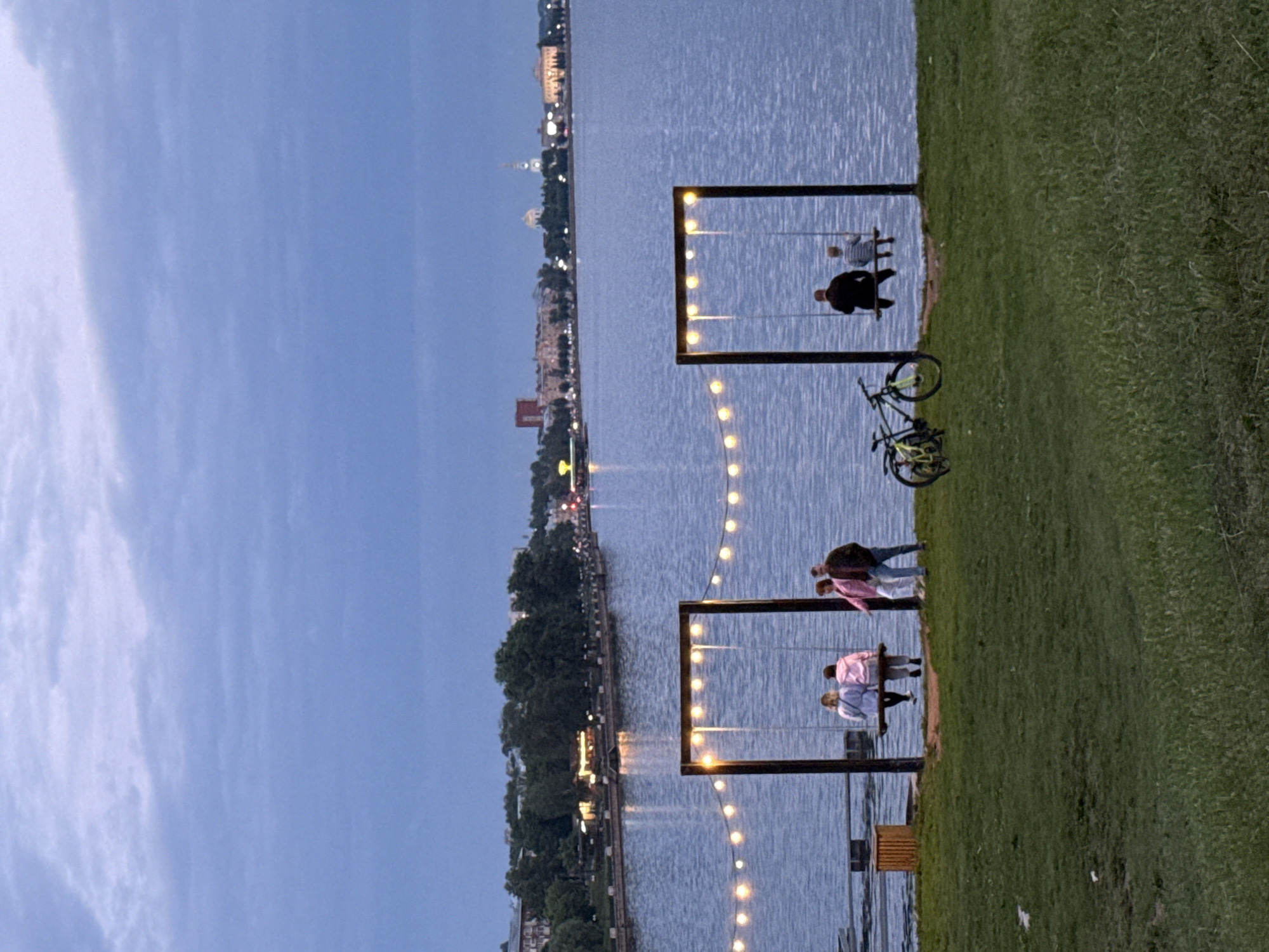 The image depicts a waterfront scene in Votkinsk, Russia, with a large body of water in the foreground and a city skyline in the background. Two prominent swings, each supported by dark wooden frames and adorned with string lights, are positioned along the water's edge. The swing on the left has two individuals, appearing to be young women, sitting on it. The swing on the right has two individuals, an adult and a child, also seated. In the grassy area between the swings and the water, a bicycle is visible. The sky is overcast with a twilight ambiance, suggesting it is early evening or late afternoon. The city skyline features buildings of various architectural styles, some illuminated, and a church spire. The overall atmosphere is tranquil and serene, with a focus on the leisure activities of the people present.