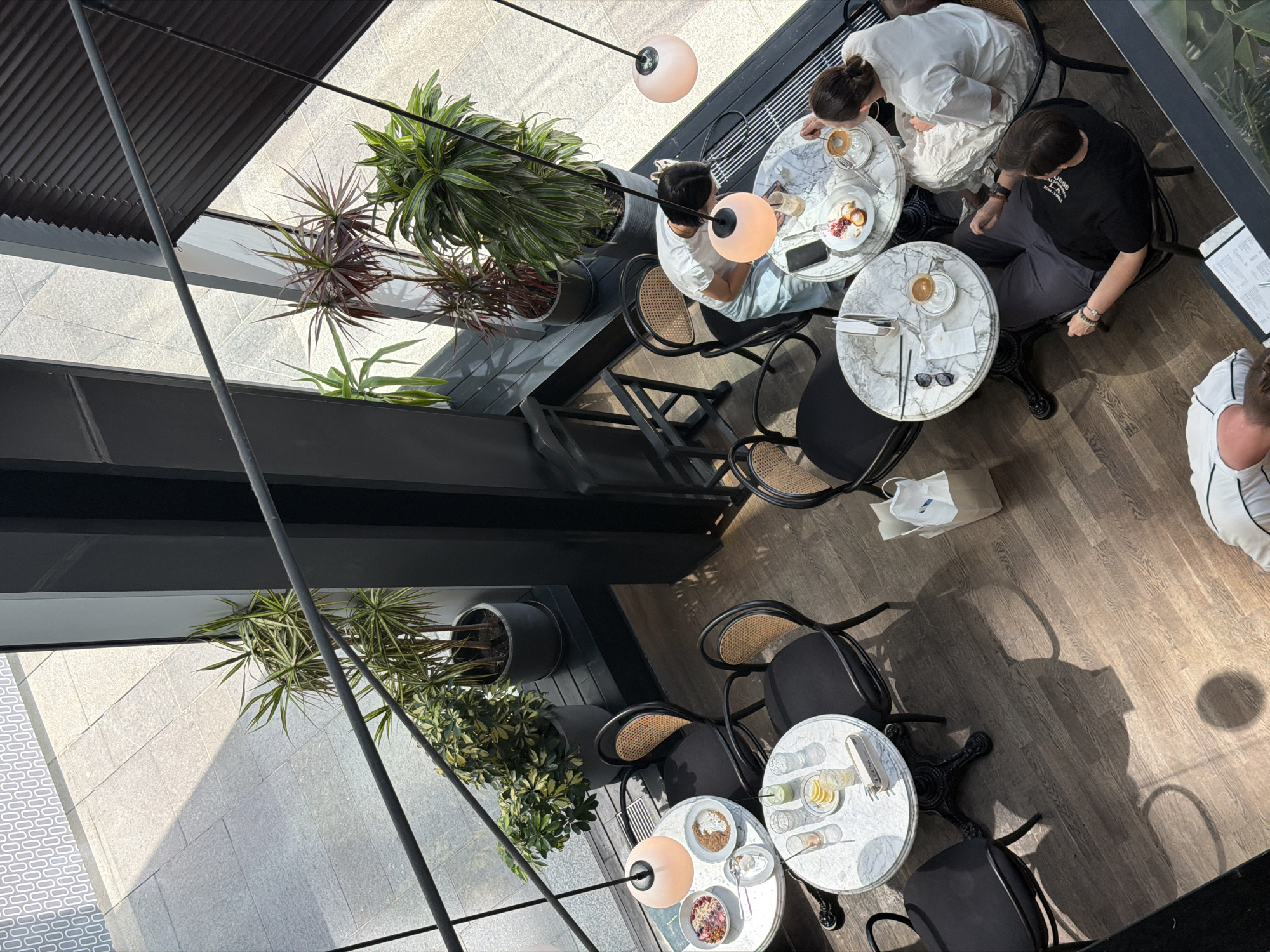 An aerial view captures a cafe interior in Almaty, Kazakhstan, where three individuals are seated at marble-topped tables. Two women, positioned at the right side of the frame, are engaged in conversation while one prepares to stir her coffee. Their attire consists of a white t-shirt and light blue pants for one woman, and a white t-shirt and white skirt for the other. On the left, a man in dark gray pants and a black t-shirt reclines in his chair, looking down. A white shopping bag rests on the floor nearby. The cafe features dark wood flooring, black chairs with wicker backs, and several potted plants adding greenery to the space. Soft pink pendant lights illuminate the tables, casting warm glows. Outside the large windows, sunlight filters through, creating distinct shadows on the tiled walkway and patterned walls, suggesting an outdoor seating area or public space adjacent to the cafe. The scene appears to be a relaxed, daytime setting.