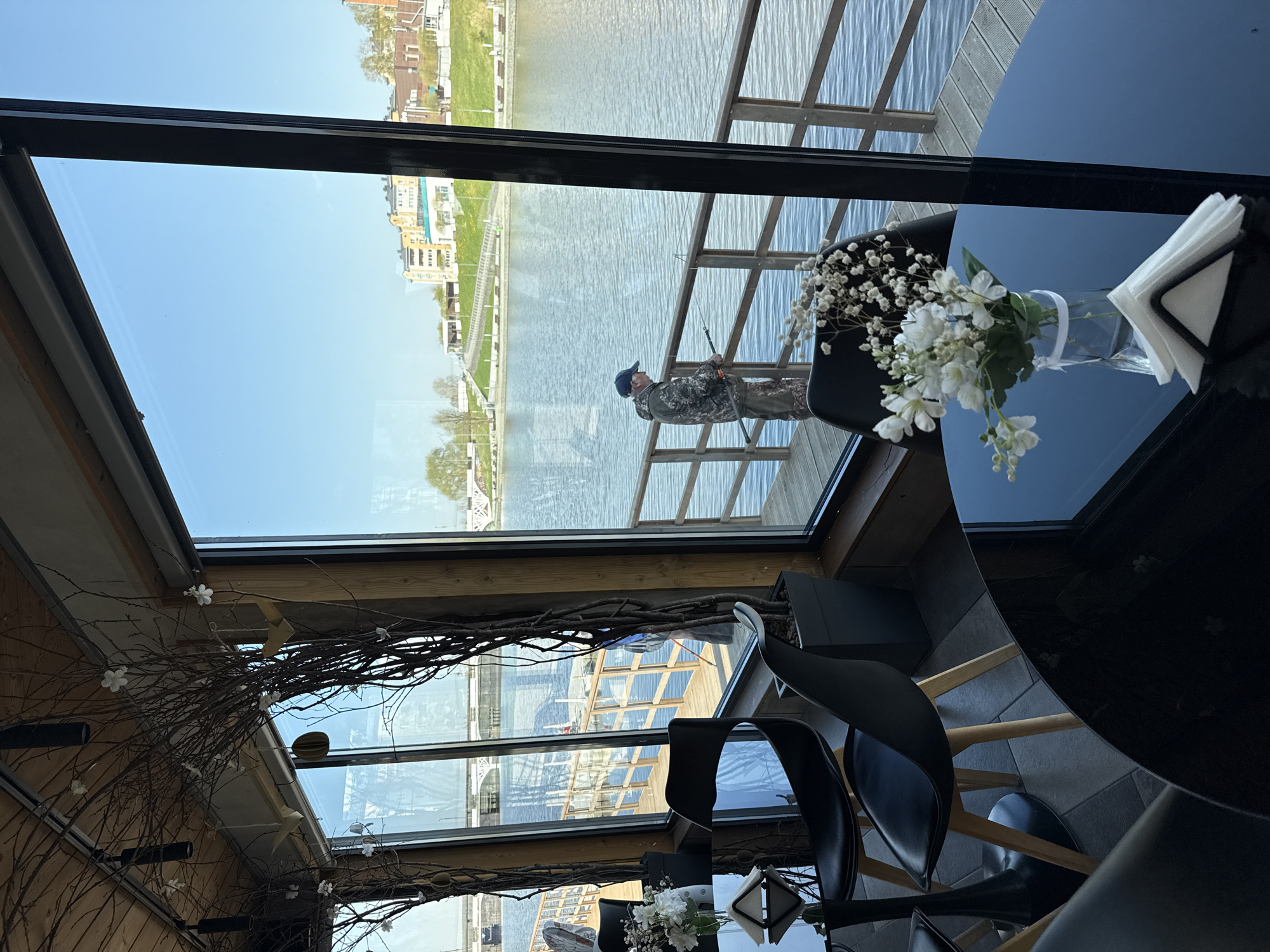 View from a restaurant, likely floating on the water, in Votkinsk, Russia. A man stands outside on a wooden deck or platform, fishing with a rod. The man is dressed in camouflage-patterned clothing and a blue cap. The restaurant interior features black chairs and tables; a vase of white flowers and napkins sit on a glossy black table. Decorative branches are affixed near the ceiling. The water reflects the cityscape and sky.