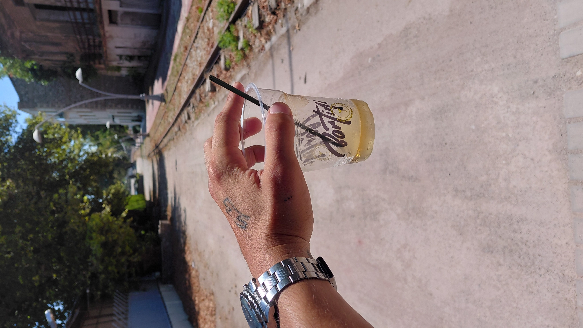 A person's hand with a silver watch on the left wrist and a tattoo on the back of the hand holds a clear plastic cup filled with a pale yellow liquid and a black straw. The cup has a logo that reads "PORTIFICI" in black lettering. The background shows a dirt path with railroad tracks running parallel to it. Buildings and trees are visible in the distance, and the scene appears to be in an outdoor urban setting. The location is the Municipal Unit of Aegio, Greece, specifically near the waterfront or a former industrial area with railroad access. The lighting suggests it is daytime.