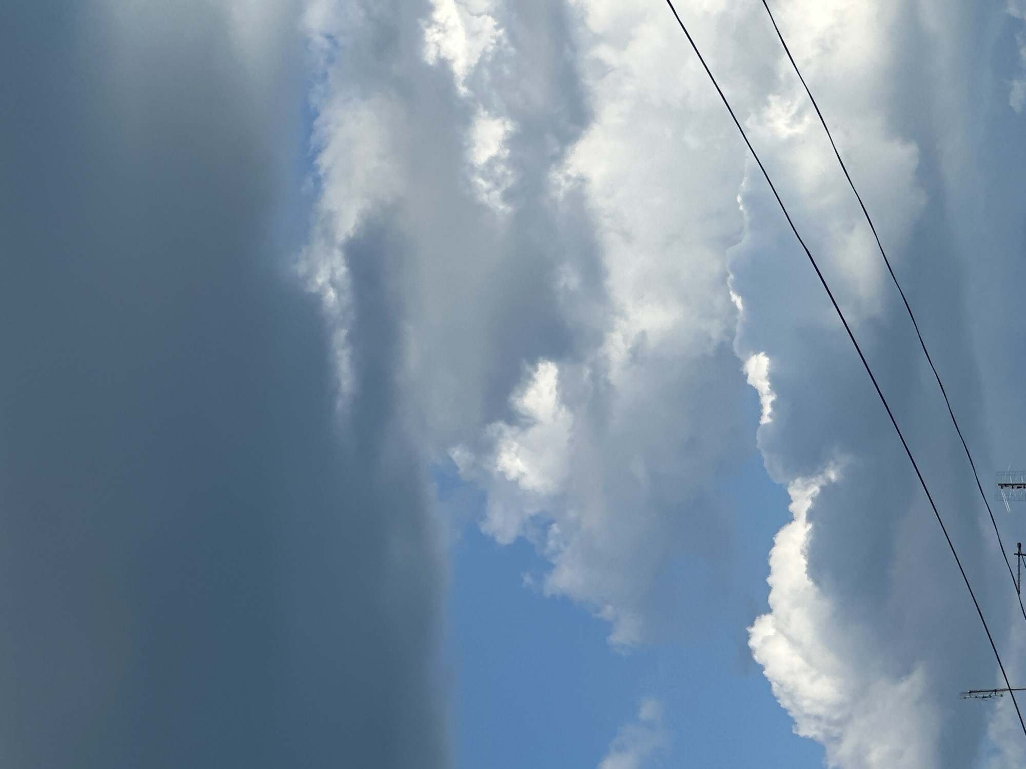 The image captures a sky filled with diverse cloud formations and utility infrastructure, situated in the city of Unknown, Unknown. The upper portion of the frame, predominantly the top left and center-right, is occupied by a dense, dark grey to bluish-grey cloud mass, suggesting heavy cloud cover. In the middle-left section, a distinct patch of clear, bright blue sky is visible, creating a strong contrast with the darker clouds. Below this, across the lower mid-ground and bottom, numerous white, fluffy cumulus clouds are present, brightly illuminated, suggesting sunlight from an unseen source. These white clouds extend to the horizon line, which is not visible.
Two prominent, dark, parallel utility wires or cables traverse the lower third of the image horizontally, angling slightly upwards from the left edge towards the right. In the bottom-left corner, a partial view of a metallic antenna or mast structure with cross-members is discernible. Another partial metallic structure, possibly another antenna or a rooftop apparatus, is visible in the bottom-right corner. There are no human figures, animals, vehicles, or specific ground-level landmarks visible in the frame. The overall environment suggests an elevated or upward-looking perspective within an urban or suburban setting, characterized by mixed weather conditions.