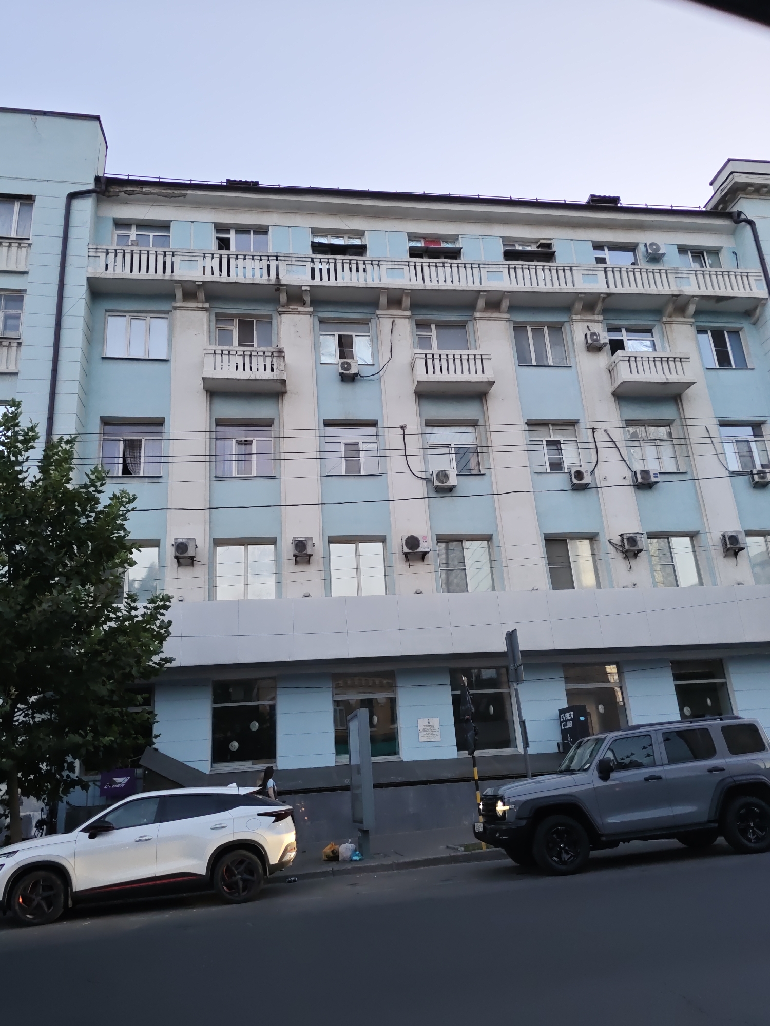 A light blue and white multi-story building with balconies and numerous air conditioning units is visible. Several windows are open, and one balcony has a red object inside. Below the building, two vehicles are parked on the street: a white SUV with red accents on its wheels and a grey SUV. A tree is to the left of the building, and power lines are strung across the facade. The sky is clear and light blue. A sign on the building reads "CYBER CLUB." The location is the city of Rostov-on-Don, Russia.
