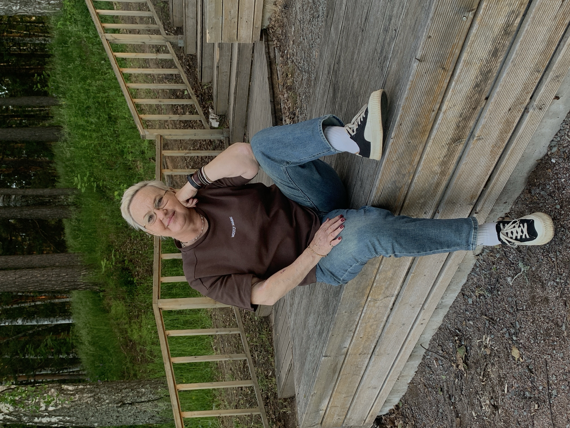 An adult female with short, light-colored hair and eyeglasses is seated on the lowest tier of a multi-level wooden structure, likely a deck or bench, in Chaikovsky, Russia. She is wearing a brown, short-sleeved t-shirt with the words "MOSTLY MAGIC" printed in light text on the chest, light blue denim jeans cuffed at the ankles, white socks, and black and off-white platform sneakers. Her right arm is bent, with her elbow resting on her right knee and her chin supported by her right hand. Her left hand rests on her left thigh. She has multiple dark-colored bracelets on her left wrist, a thin gold-toned bracelet on her right wrist, and a ring on her left ring finger. Her fingernails are painted red. A light-colored beaded necklace is visible around her neck. The wooden structure she sits on is weathered. In the background, a wooden railing extends laterally and then forms a balustrade for a set of wooden stairs ascending to the right. The environment behind the railing and stairs is a dense forest with various trees, including white-barked birches and darker-trunked conifers, and lush green undergrowth. The ground immediately in front of the wooden structure is covered with gravel and scattered dry leaves.