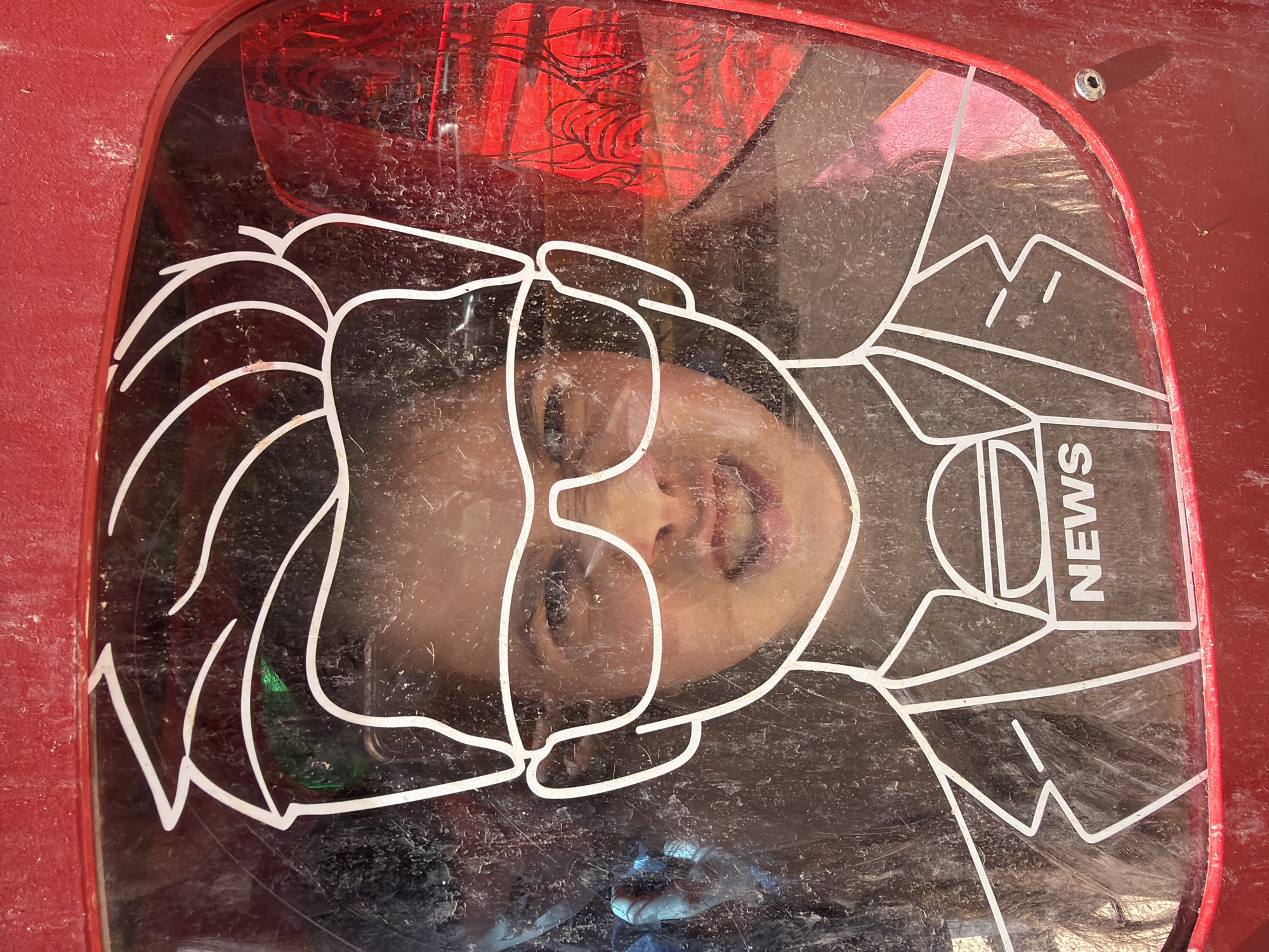 A low-angle, medium shot shows a child's face seen through a display case or window, with a white line drawing of a news reporter overlaid. The reporter figure includes a microphone labeled "NEWS" and outlines of clothing. The child is looking up at the camera, and their face is distorted by the glass or plastic of the display. The case appears dirty, with smudges and dust obscuring the view. The display case is red and appears to be mounted on a larger red structure. Based on the drawing of a news reporter, this appears to be located in Yerevan, Armenia.
