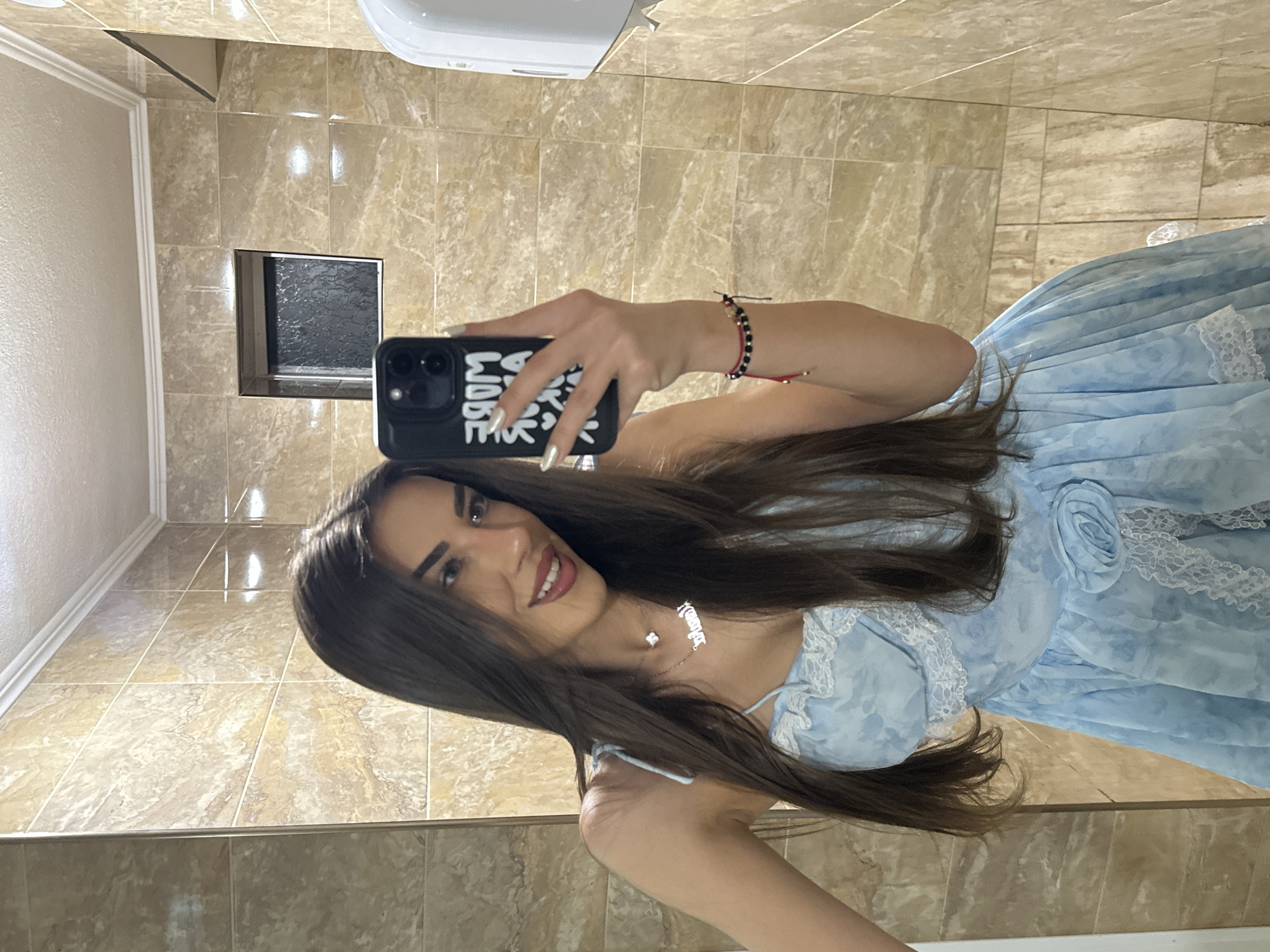 A young woman is taking a mirror selfie in a tiled bathroom. She has long dark hair and is wearing a light blue dress with lace trim. Her phone case has white text on it. She is also wearing a necklace and bracelets. The bathroom has beige marble tiles on the walls and a white ceiling.