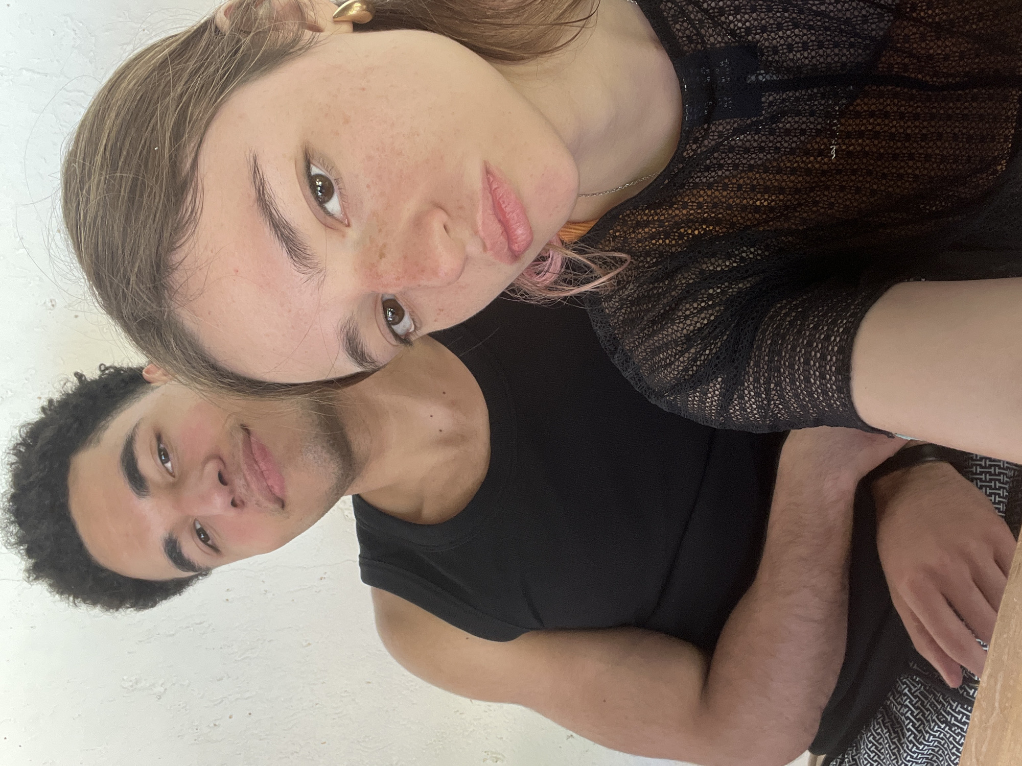 A tilted medium shot shows a light-skinned woman and a dark-skinned man in an embrace. The woman, positioned at the top of the frame, looks directly at the camera with a slight pout. She has brown hair with some pink streaks, freckles, and is wearing a black mesh top. The man, located at the bottom of the frame, is smiling slightly. He has short, curly black hair and wears a black tank top. They are indoors with a white wall visible in the background. The location is unknown.