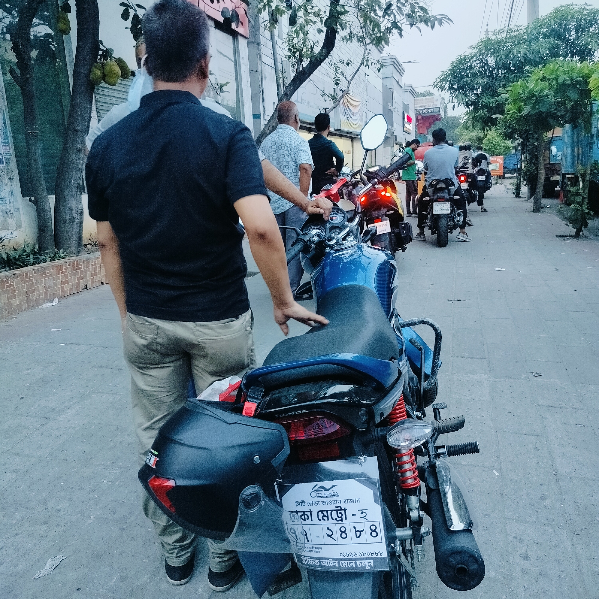 A street scene in Dhaka, Bangladesh, features a line of motorcycles and several people, most of whom are facing away from the viewer. A man in a black shirt and khaki pants stands beside a blue motorcycle, his hand resting on the seat. The motorcycle's license plate reads "Dhaka Metro - H 77-2484." The license plate also shows that the motorcycle has been sold by CITY HONDA which is at Kawran Bazar. The license plate also has a contact phone number 01819 170444. The adjacent motorcycles are similarly oriented. Other people stand nearby, appearing to be affiliated with the motorcycles. The backdrop consists of storefronts, trees, and utility poles. The scene seems to be in daylight, with a mixture of natural light and shadows.