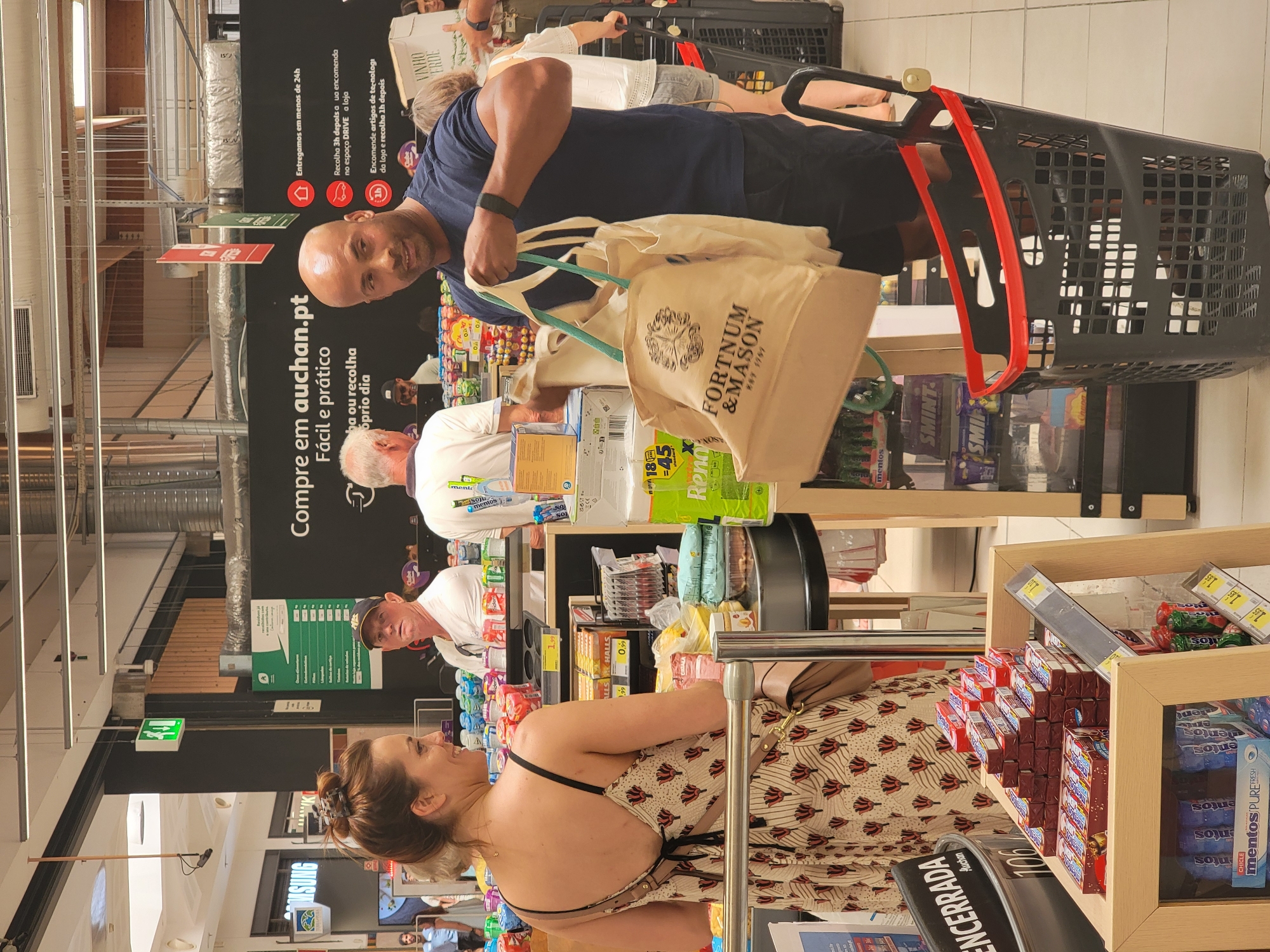 The image depicts a busy scene inside a supermarket in Faro, Portugal. In the foreground, a man of African descent wearing a dark blue t-shirt and black shorts is holding two reusable shopping bags, one of which prominently displays the logo "FORTNUM & MASON EST. 1707". He is looking towards the camera with a slight smile. To his left, a woman with brown hair tied up is wearing a patterned dress and looking to her left. Behind them, other shoppers and displays of products are visible. The environment includes bright fluorescent lighting and shelving stocked with various grocery items. A black shopping cart with a red trim is positioned in the lower right portion of the frame. The store appears to be an Auchan supermarket, suggested by signage in the background.