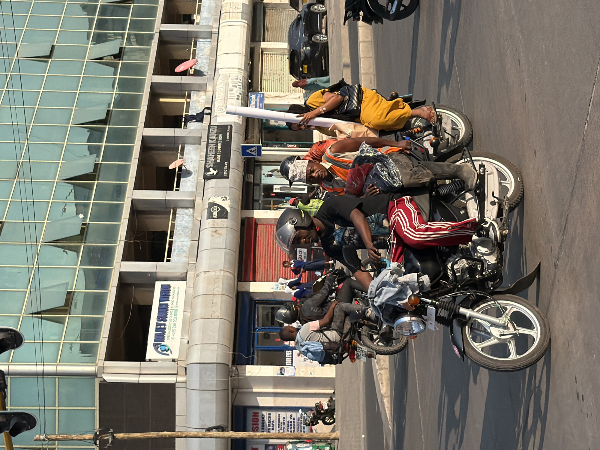 This image captures a street scene in Dar es Salaam, Tanzania, featuring several motorcycles and pedestrians in front of a modern building with a glass facade. In the foreground, a man wearing a helmet and red and white striped pants rides a motorcycle. He is accompanied by a second person on the motorcycle, also wearing a helmet, and a third person in the back, holding a large white cylindrical object. Another motorcycle is visible behind them, carrying two passengers. Further in the background, pedestrians are seen walking on the sidewalk, and a black SUV is parked on the street. The building in the background displays various signs, including "MAJEY SMART VISION" and "SHAHEEN KANZU MADE TO PERFECTION." The lighting suggests it is daytime, possibly late afternoon, with long shadows cast on the road.