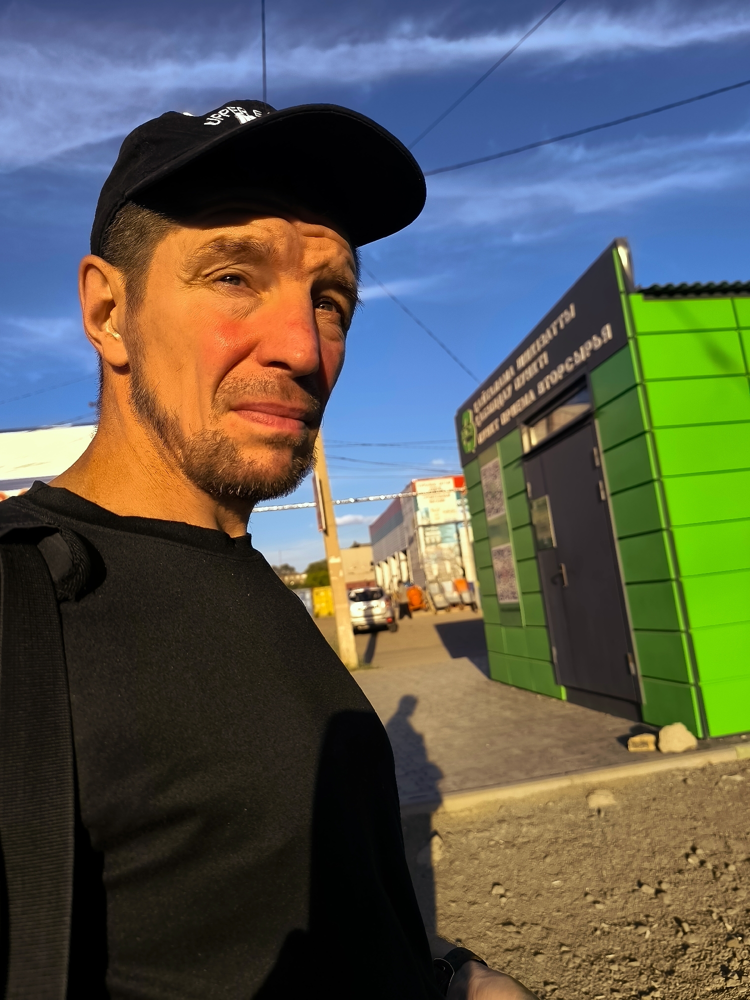 A medium shot captured from a slightly low angle shows a man in his late 30s or early 40s with a light beard, wearing a black t-shirt and a black baseball cap. He is looking towards the camera with a neutral expression. The man is positioned on the left side of the frame, with his face illuminated by warm, late afternoon sunlight.
Behind him and to the right, a bright green, modern-looking building with dark gray doors and windows is visible. The building has signage with text in Cyrillic script. The architectural style of the building and the surrounding environment, including the paved walkway and a glimpse of a street with other buildings and vehicles in the background, suggest an urban setting. A shadow of the man is cast on the ground to his left. The sky above is a vibrant blue with scattered clouds. The overall impression is a candid outdoor portrait in a public space within Qarağandı city administration, Kazakhstan.